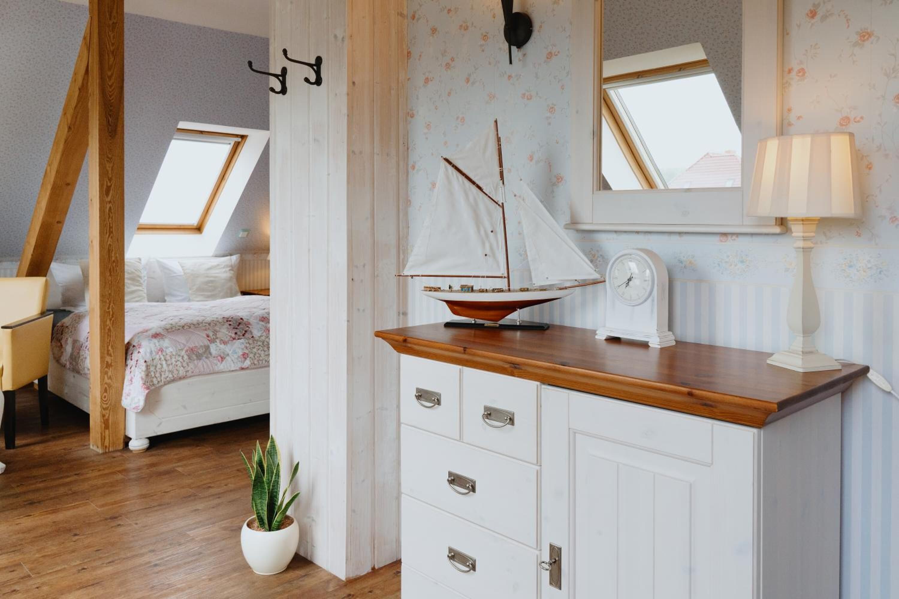 Un dormitorio con techo inclinado, una cama, un aparador, un modelo de velero, una planta en maceta y una ventana con vistas.