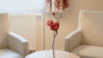 A round wooden table with a white lampshade and a vase with red flowers. Two white armchairs are positioned on either side of the table.