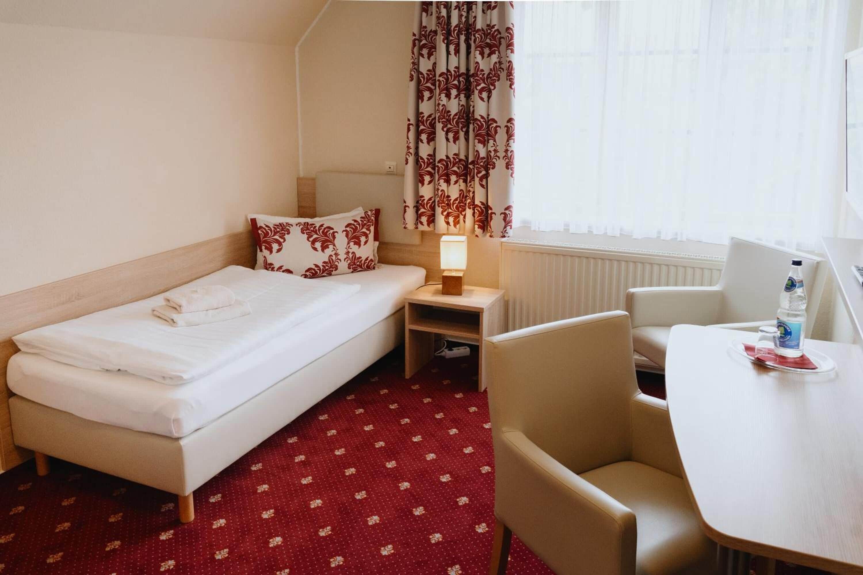 Ein Hotelzimmer mit einem Einzelbett, einem kleinen Schreibtisch, einem Sessel, einem Tisch und einem Vorhangfenster.