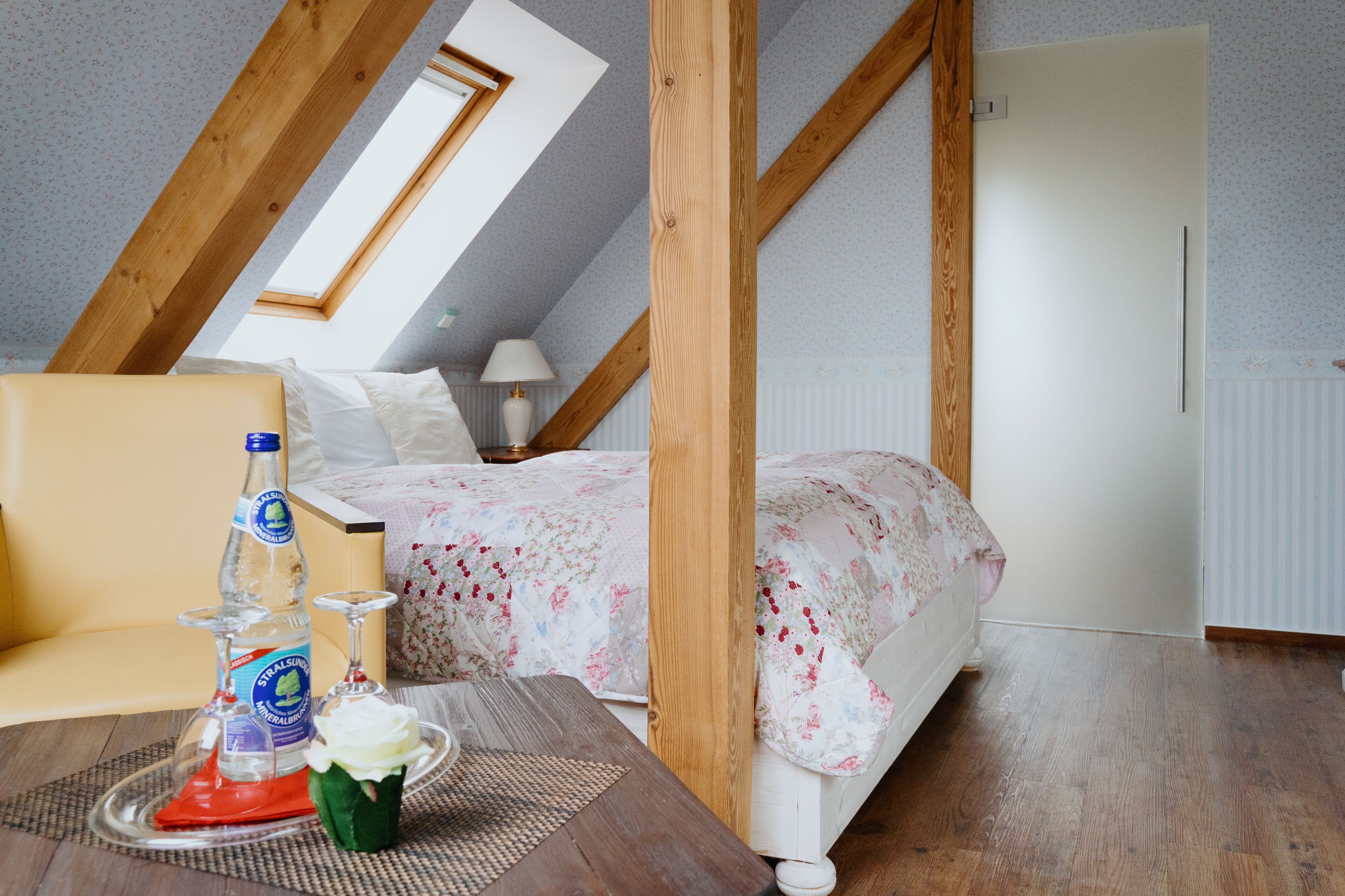 A bedroom with a sloped ceiling, a bed with floral bedding, a bedside table with a bottle and glasses, and a window with a view of the sky.