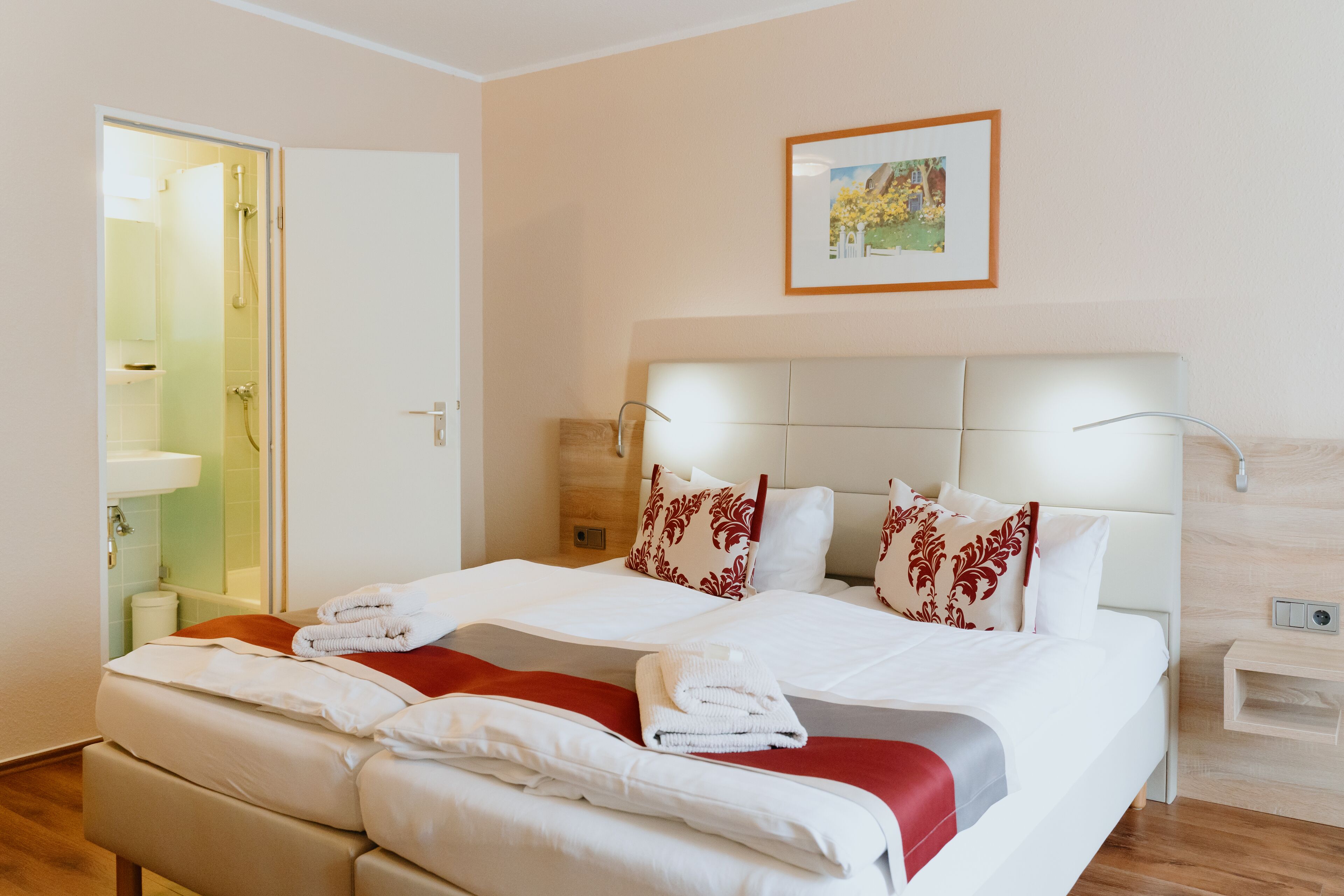 A hotel room with a large bed, a bedside table, a nightstand, a headboard, a wall-mounted picture, and a bathroom with a shower and sink.