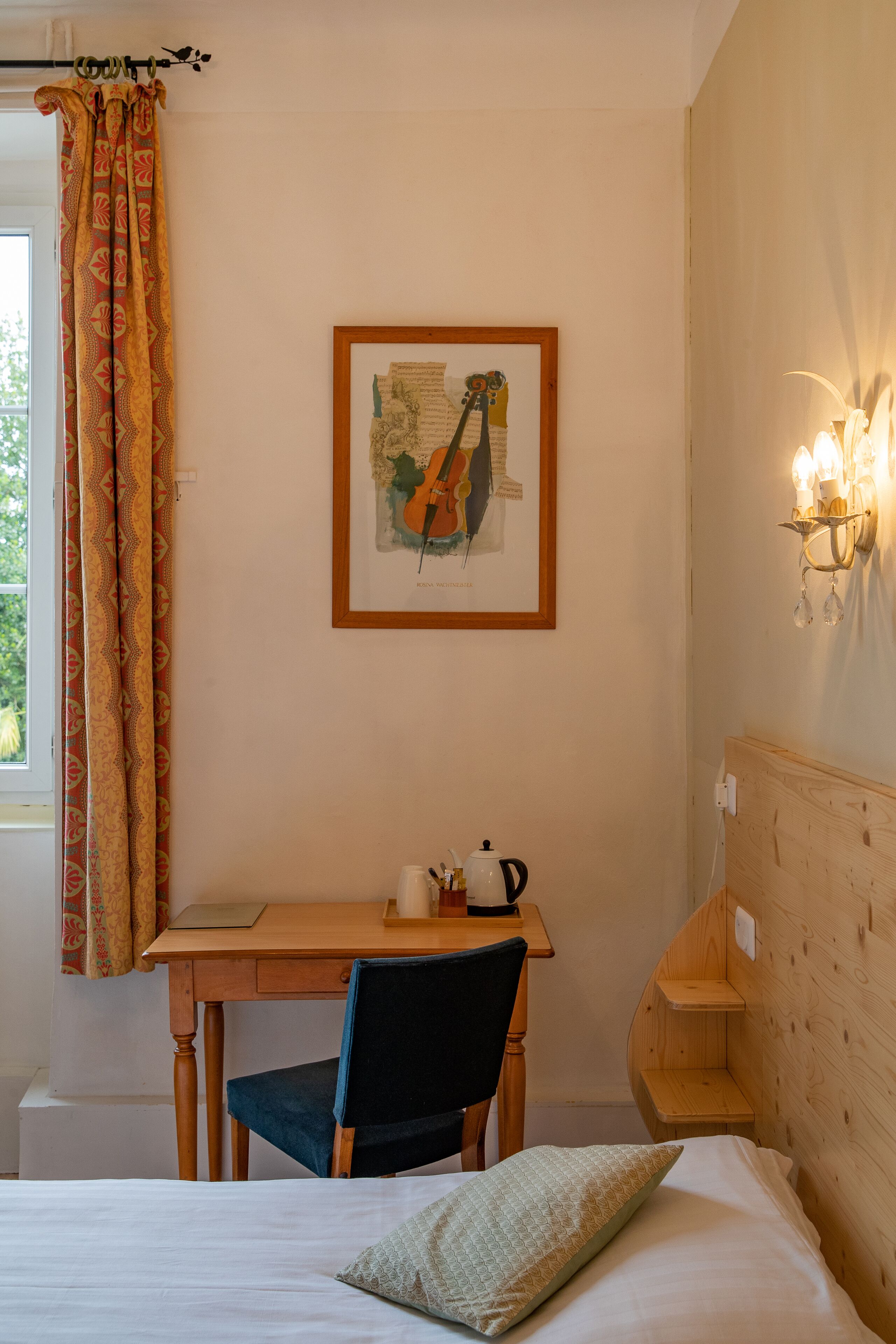 A room with a bed, a desk with a chair, a framed map on the wall, and a wall-mounted light fixture.