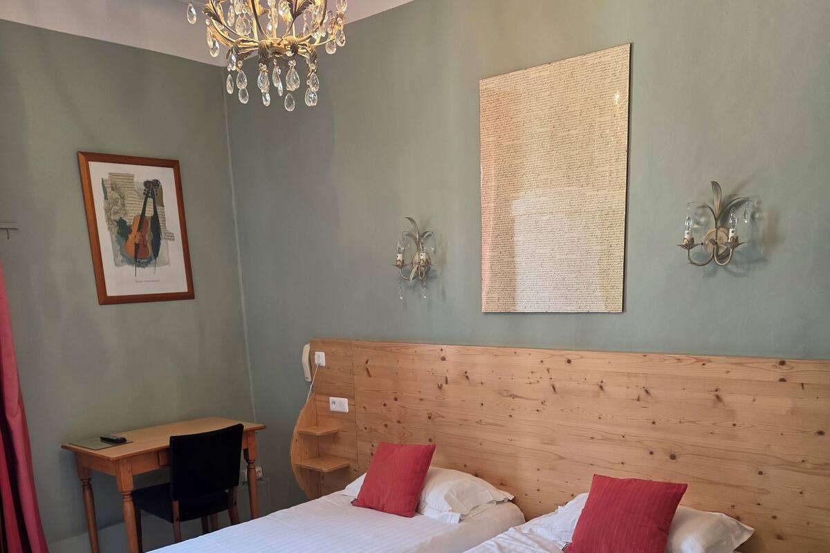 A double bed with white linens and red pillows, a wooden headboard, a desk with a chair, a chandelier, and a framed picture on the wall.
