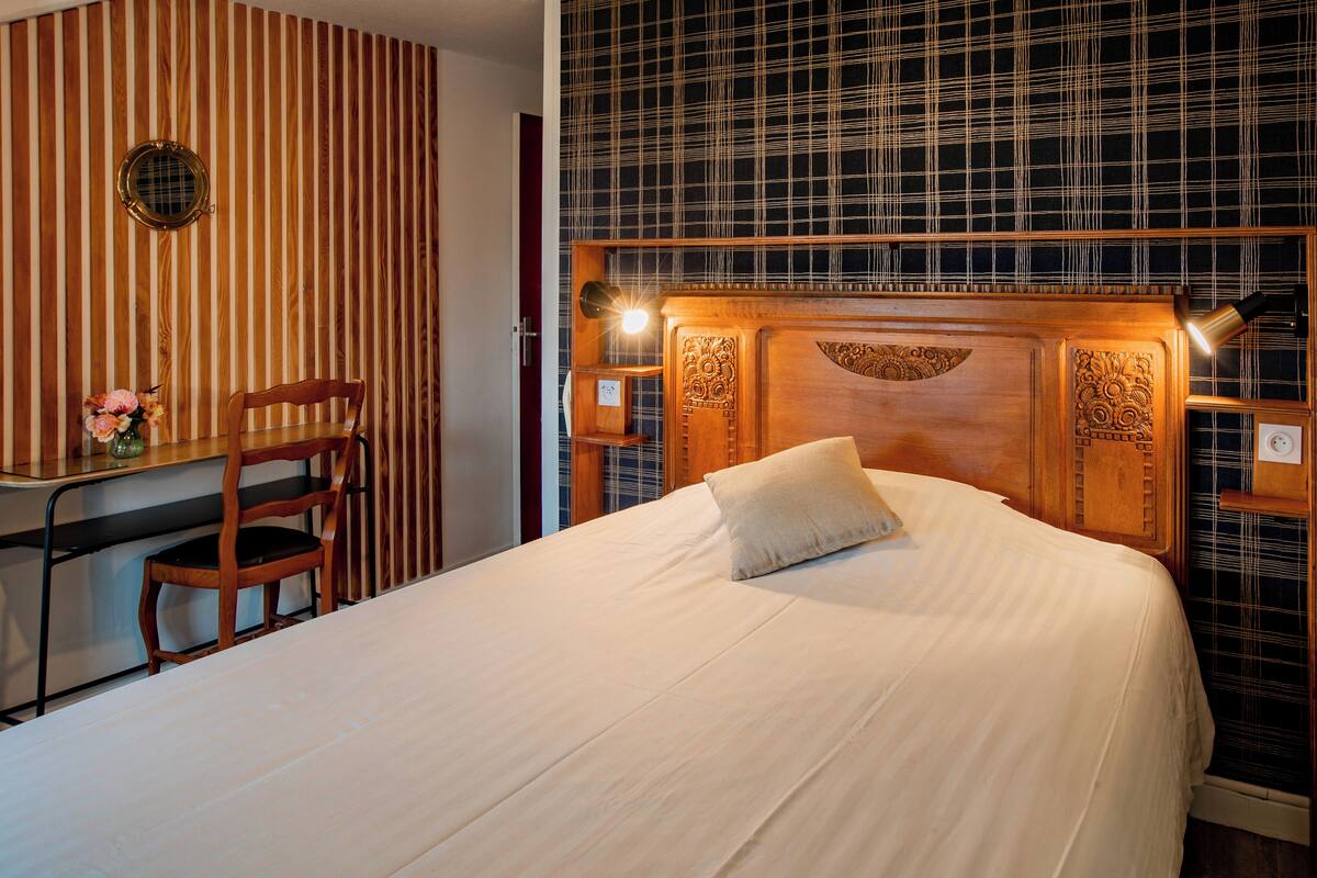 A hotel room with a wooden headboard, a bed with a white bedspread, a wooden desk with a chair, and a wall with vertical wooden slats.