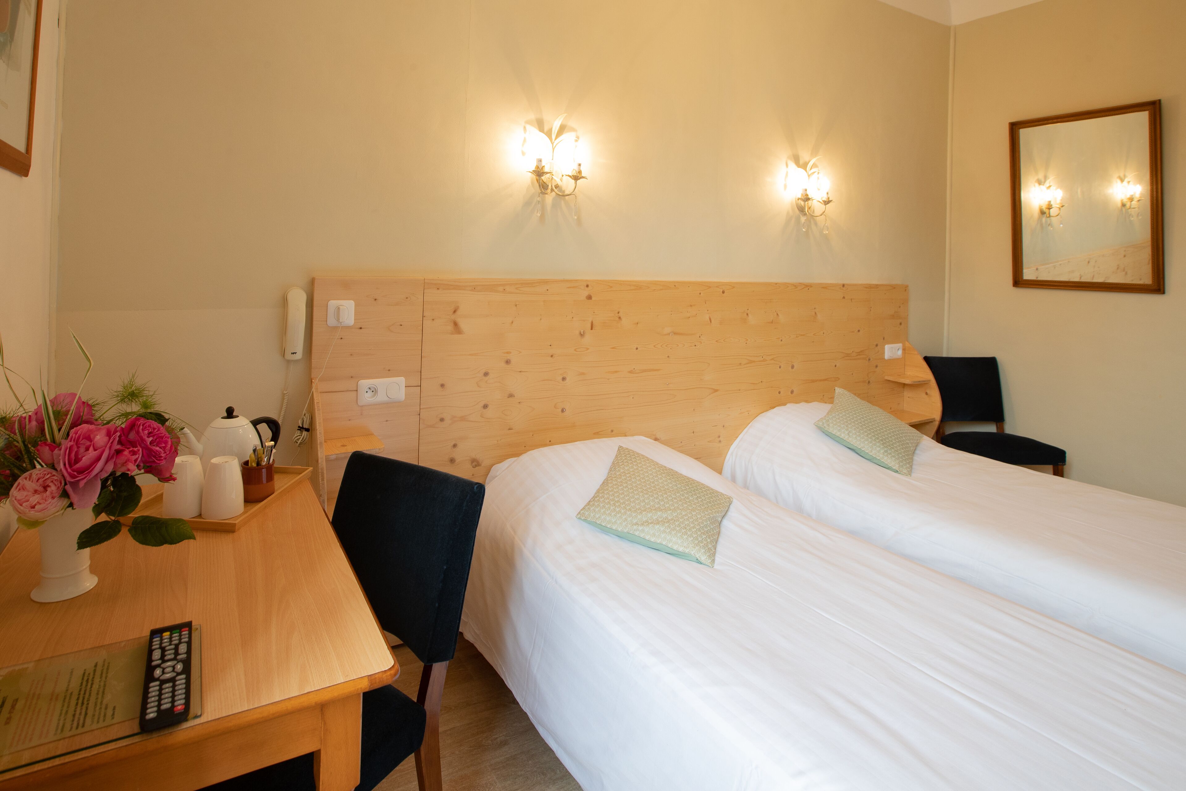 Una habitación de hotel con dos camas individuales, un cabecero de madera, un escritorio con un control remoto, una silla y un jarrón con flores.