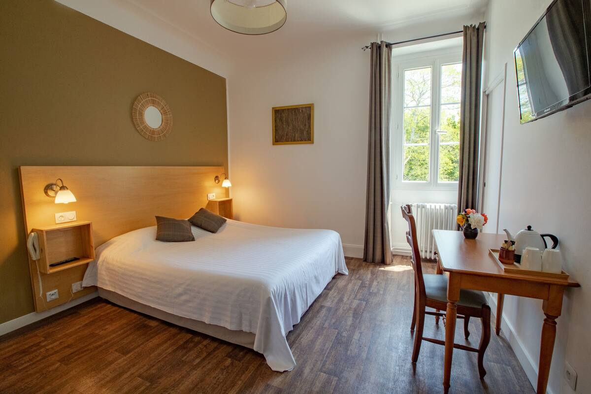 A hotel room with a bed, bedside lamps, a desk with a chair, a window with curtains, and a television on the wall.