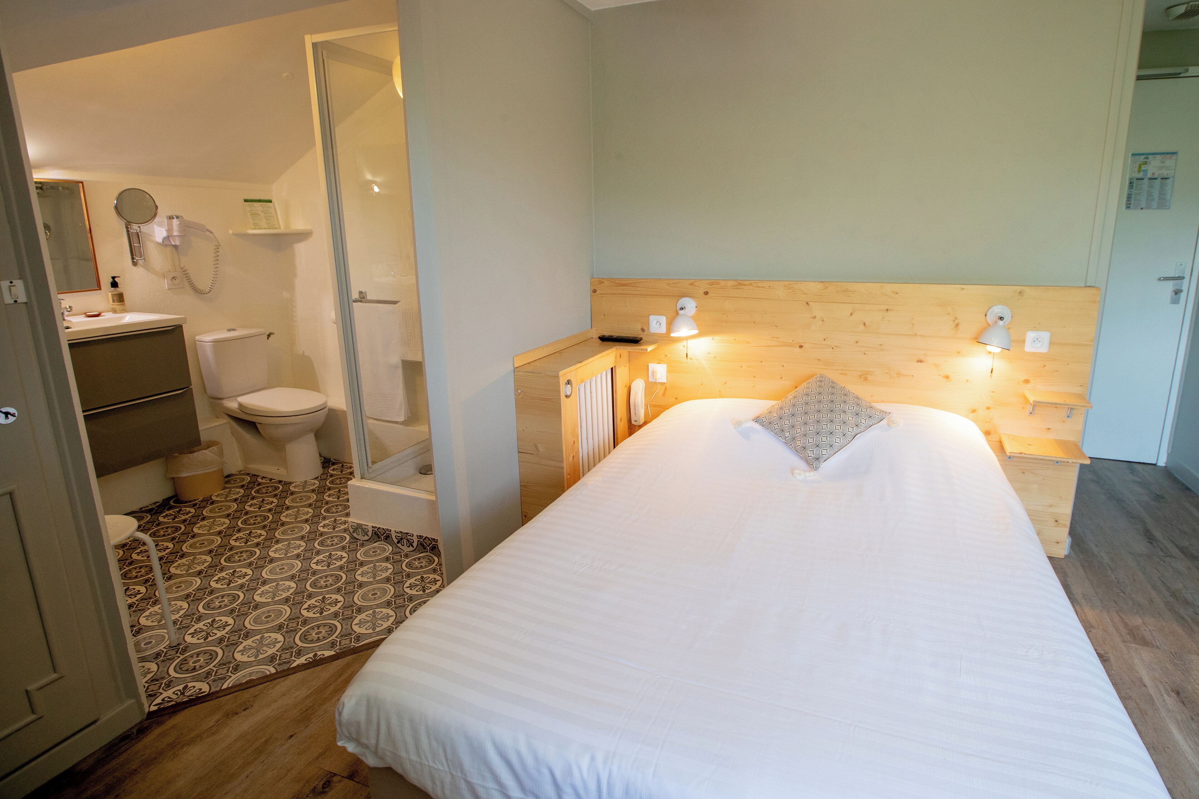 A bedroom with a bed, a wooden headboard, a bedside table with a lamp, a small shelf, and a patterned rug.