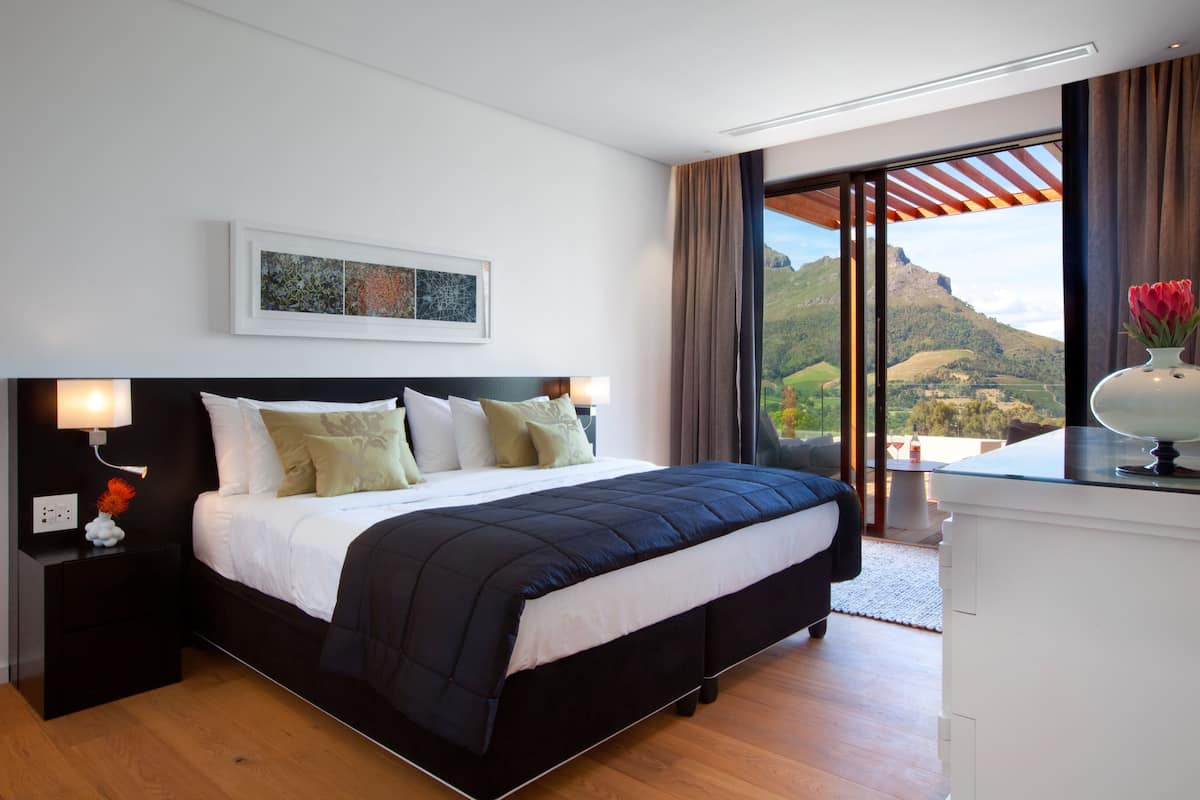 Una habitación de hotel moderna con una cama grande, una mesita de noche con lámpara, un cuadro en la pared y vistas a un paisaje montañoso a través de un ventanal corredero.