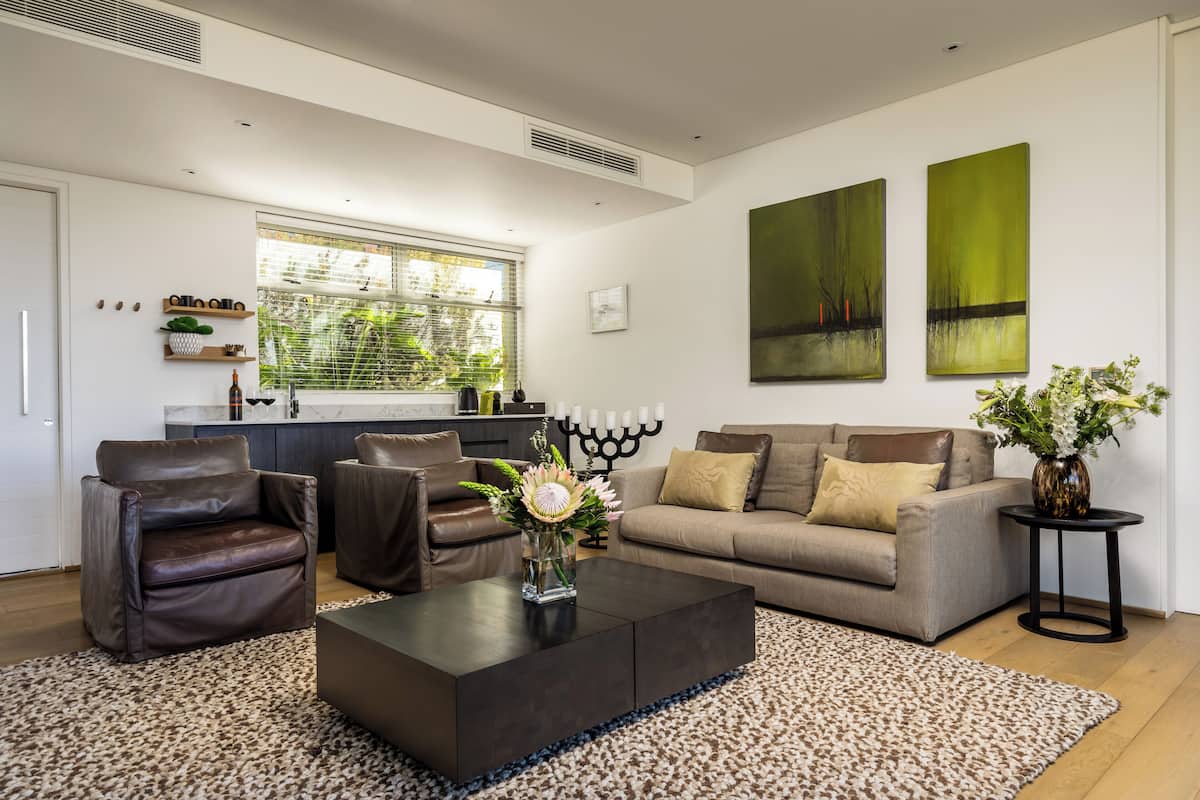 Una sala moderna con un sofá, dos sillones, una mesa de centro y un jarrón de flores. Hay una ventana con persianas, una cocina al fondo y cuadros abstractos en la pared.