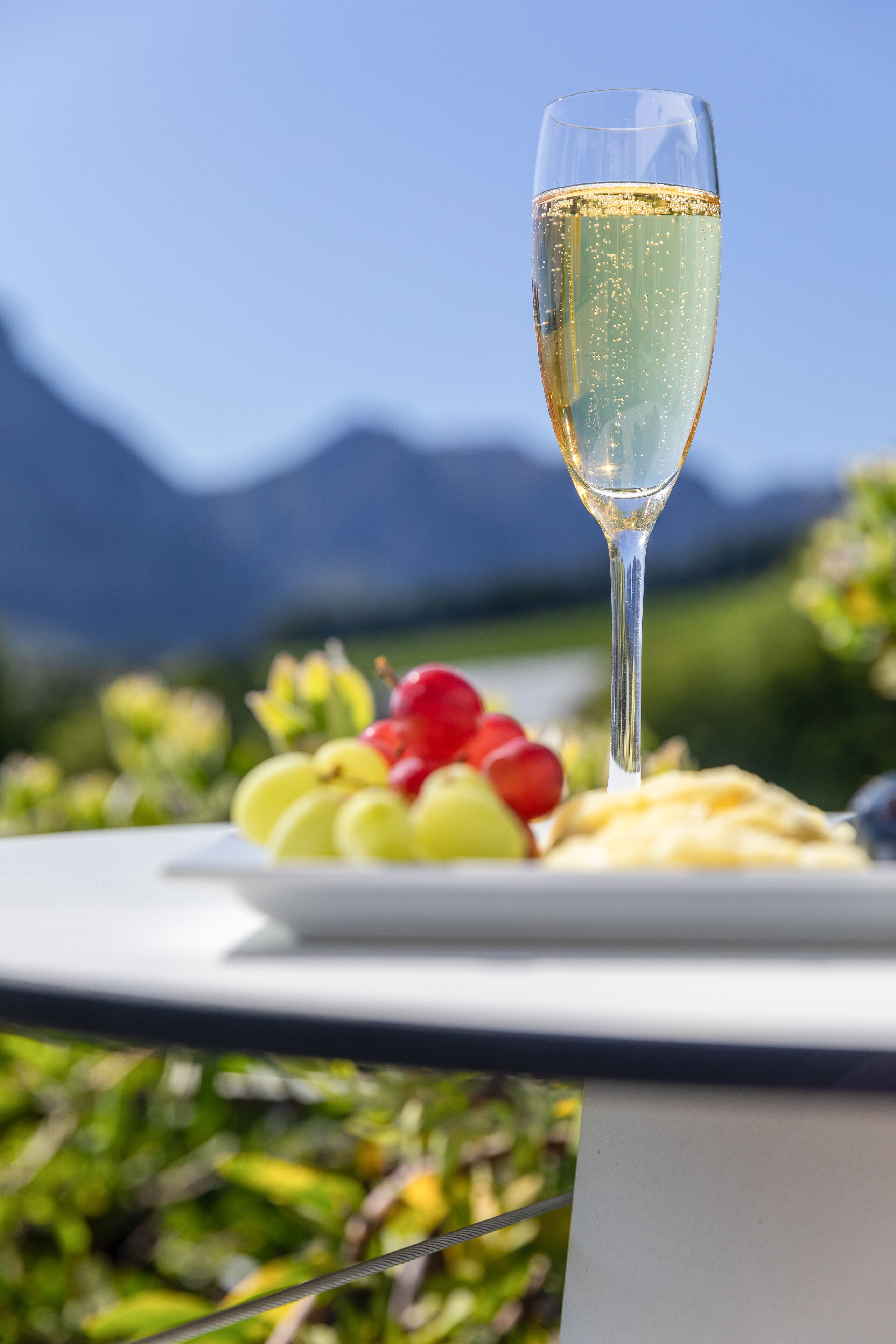 A glass of champagne on a table with a plate of grapes and cheese, set against a mountainous backdrop.