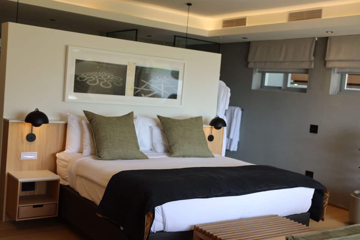Un dormitorio moderno con una cama grande, dos mesitas de noche, una lámpara auxiliar y una manta negra, además de una obra enmarcada sobre la cama.