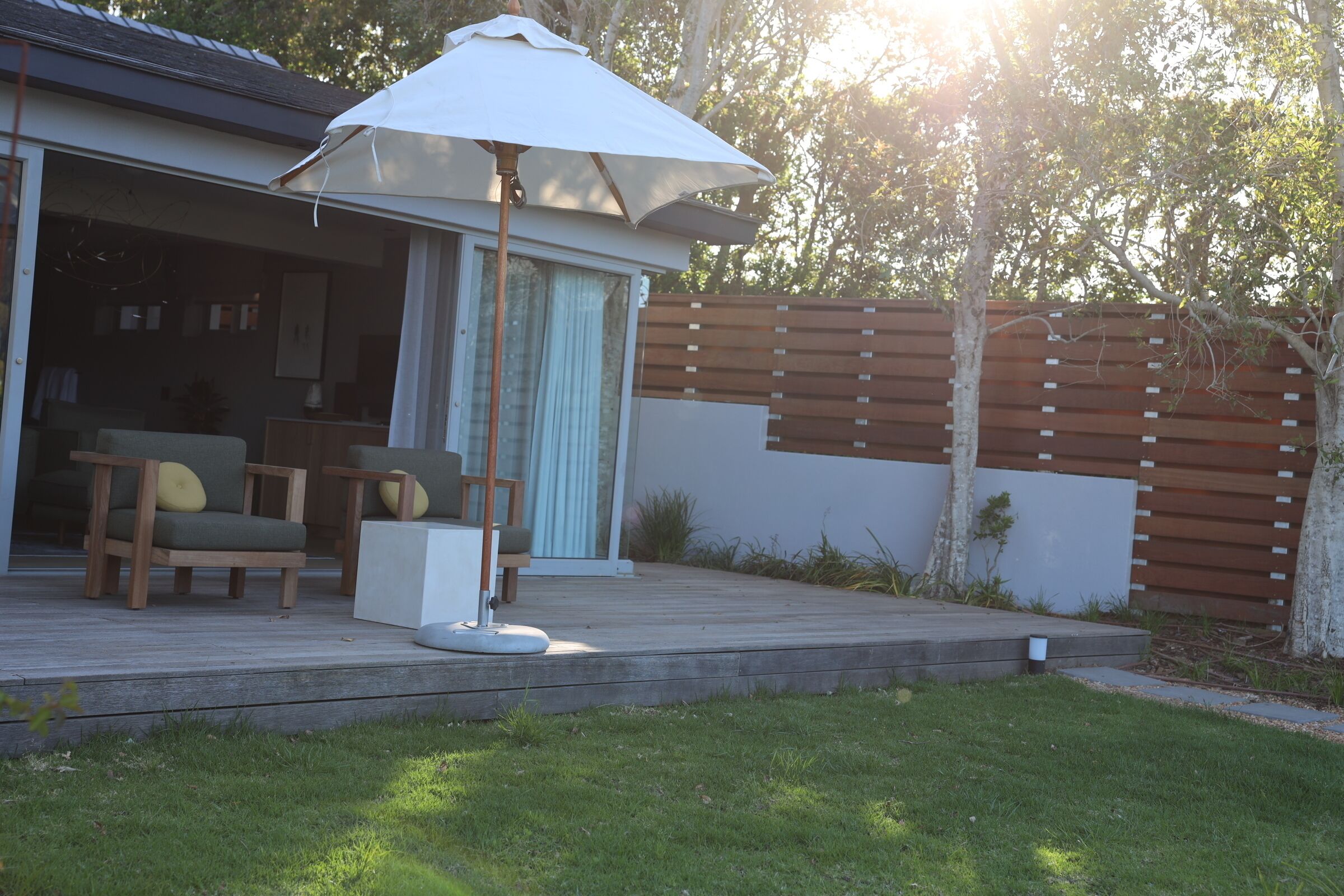 A modern outdoor deck with a white umbrella, wooden chairs, and a glass door leading to a house.