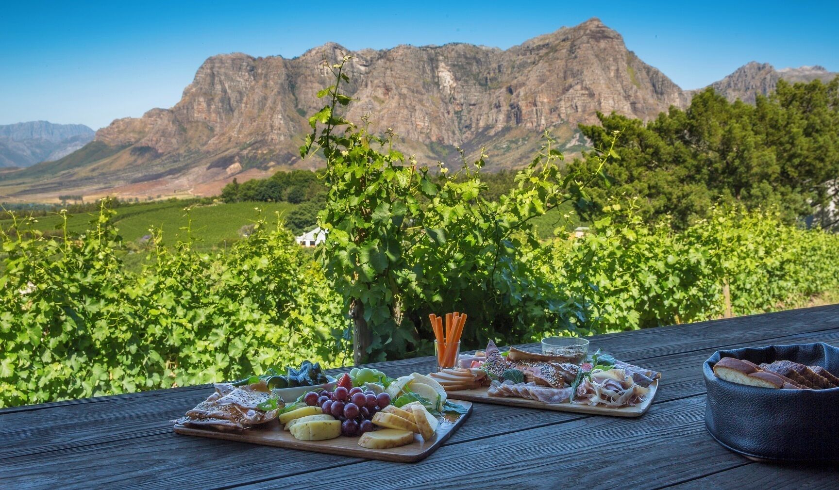 A picnic setup with various cheeses, fruits, and meats on a wooden table with a vineyard and mountains in the background.