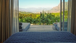 A bedroom with a large bed, a view of a vineyard, and a wooden deck. - Clouds Estate (Stellenbosch)