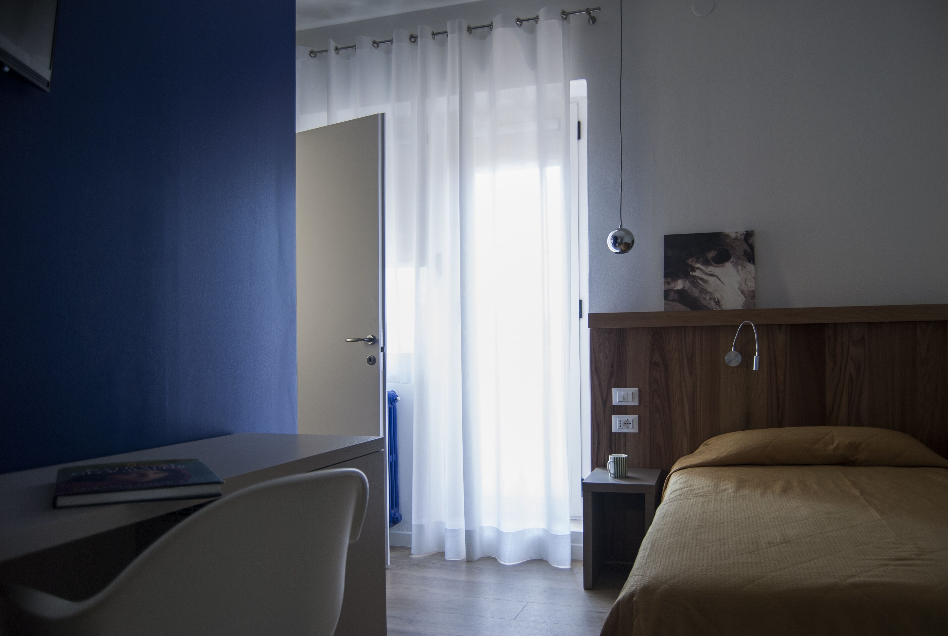 A bedroom with a bed, a desk, a chair, a bedside table, and a window with curtains.