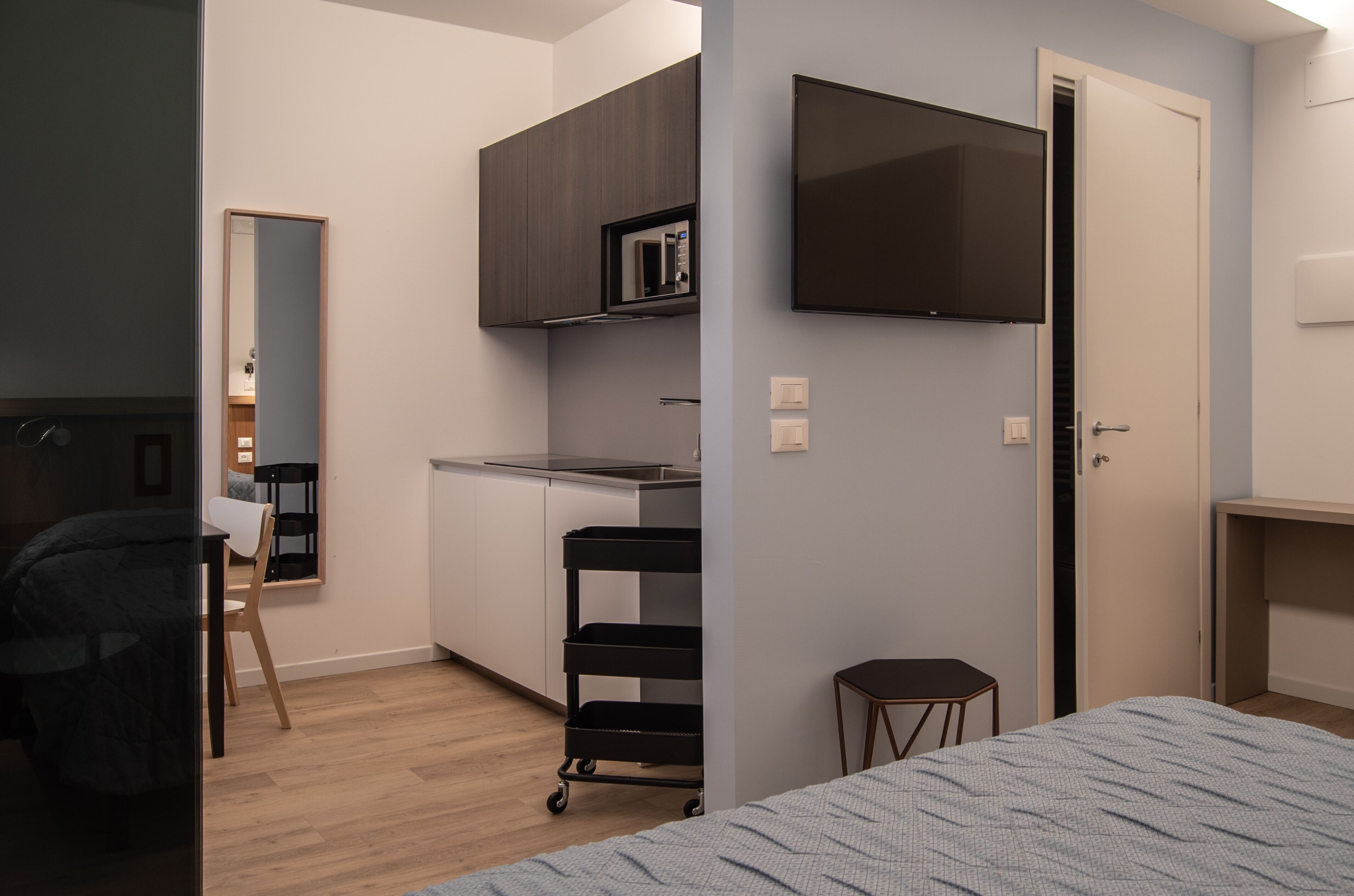 A compact hotel room with a bed, a small kitchenette, a TV mounted on the wall, and a door leading to another room.