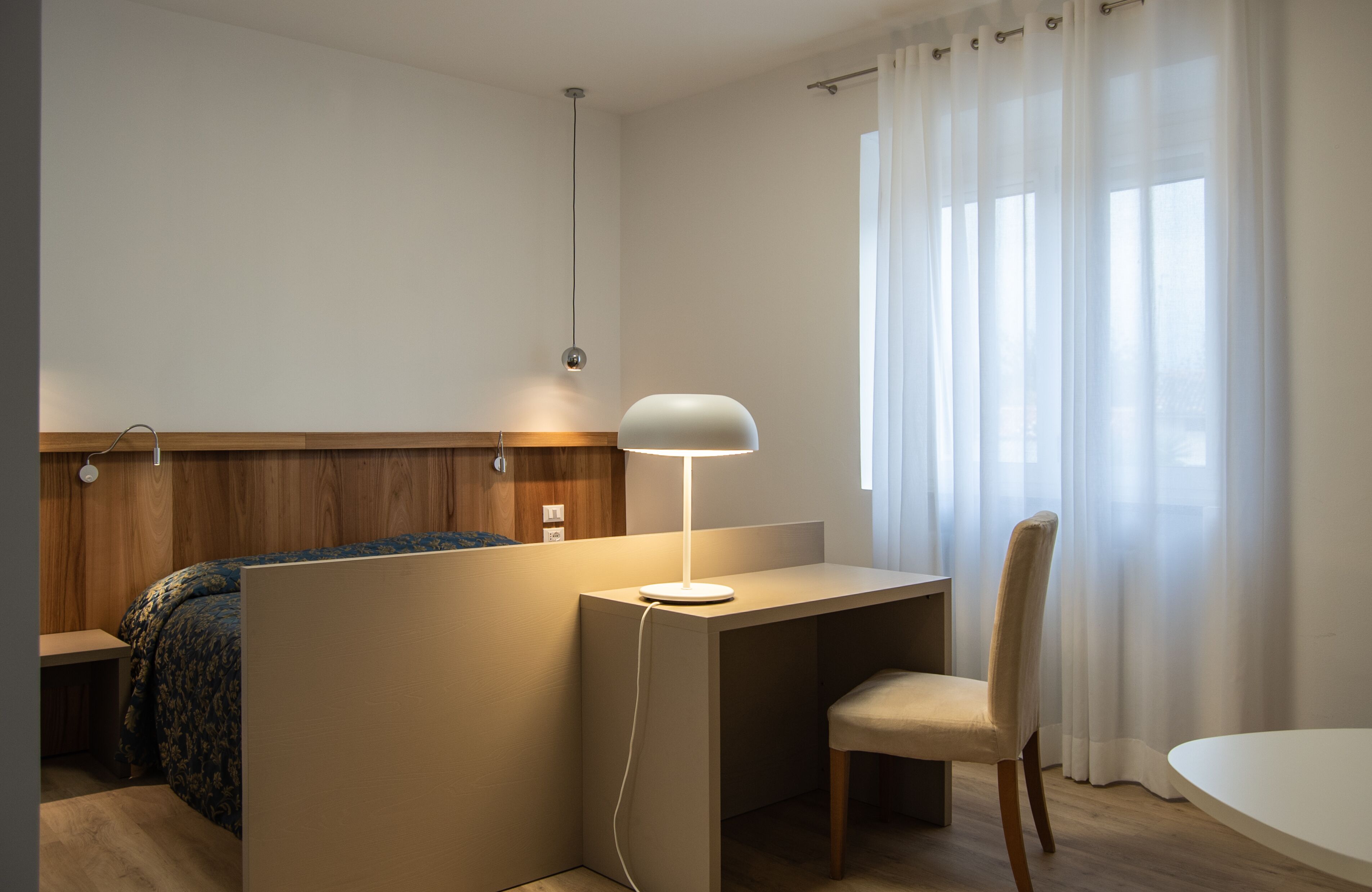 Ein Hotelzimmer mit Bett, Schreibtisch mit Lampe, Stuhl und einem fenster mit Vorhängen.