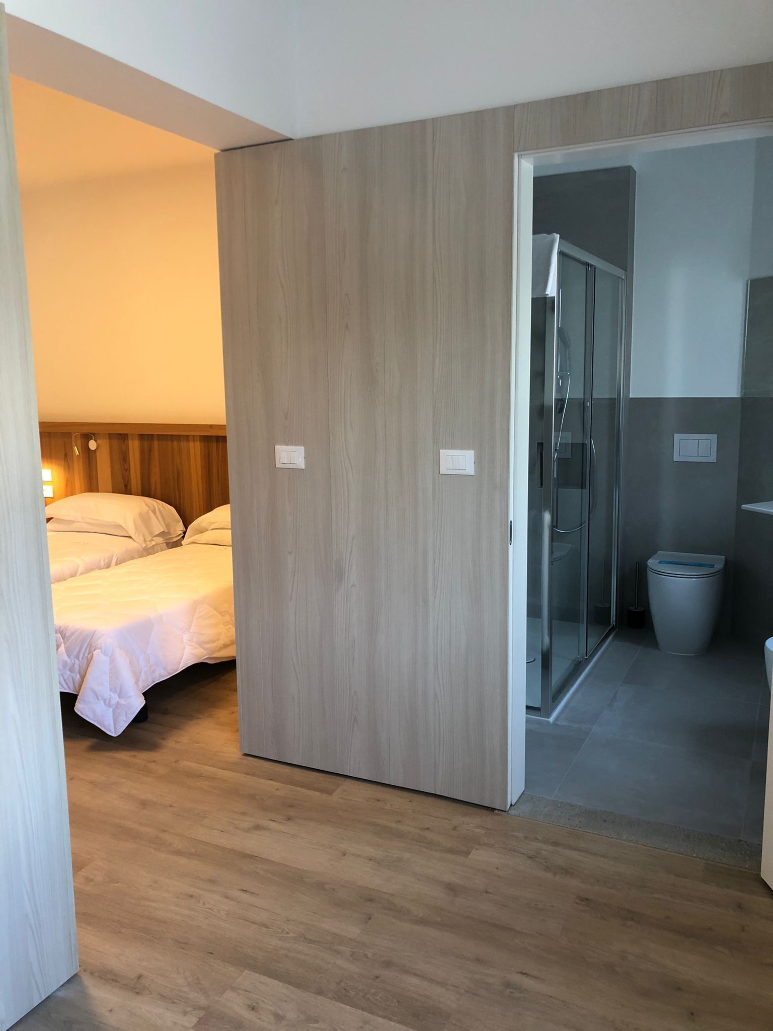 A hotel room with a wooden floor, a bed with white bedding, a glass shower enclosure, and a toilet.