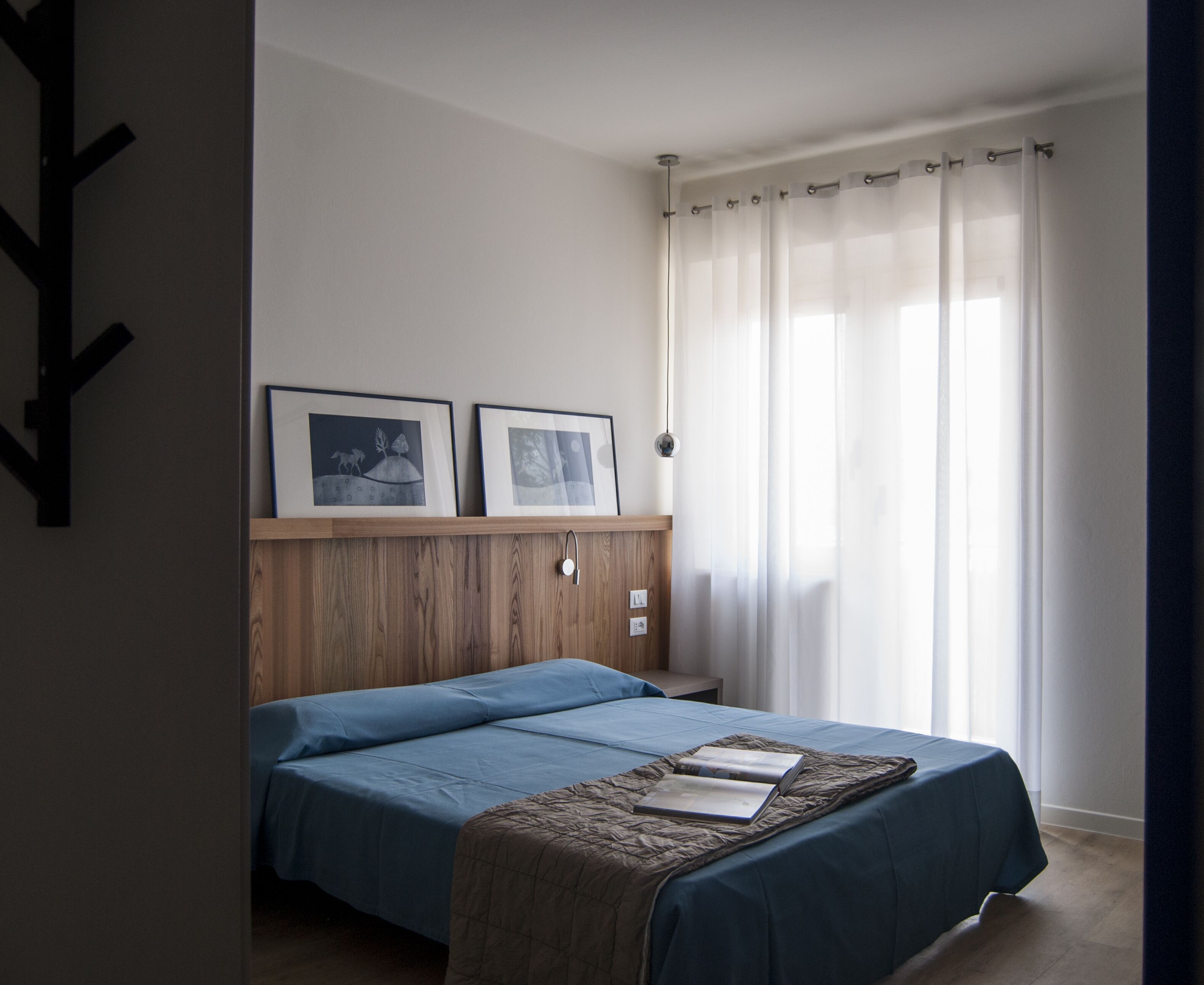 A bedroom with a bed, two framed pictures on the wall, a wooden headboard, and a window with curtains.