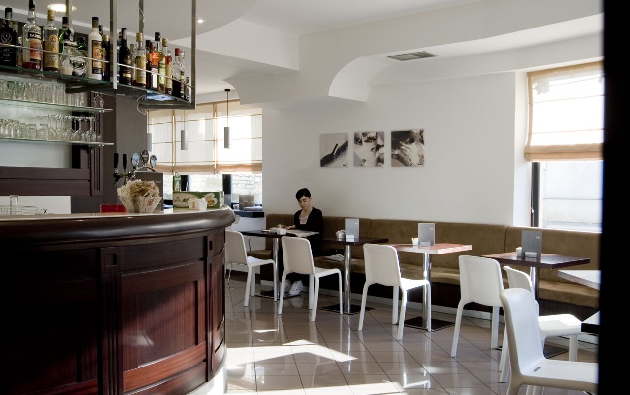 Zona de bar moderna con una variedad de botellas de licores y vasos, sillas blancas y un espacio de estar con un sofá y mesas.
