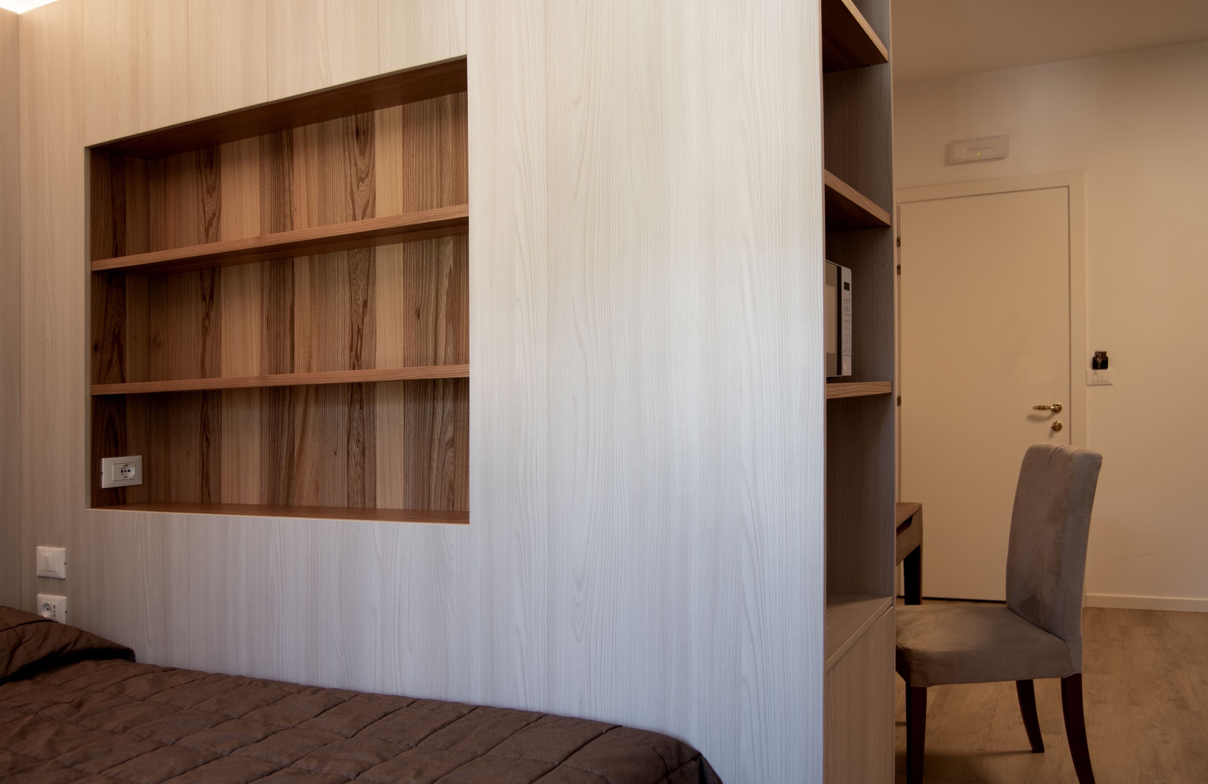 A bedroom with a bed, a wooden bookshelf, a grey chair, and a door.