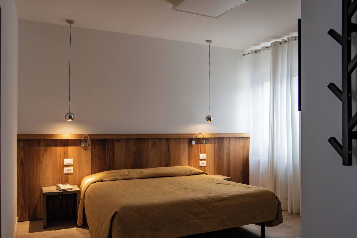 A modern hotel room with a wooden headboard, a single bed, bedside tables with lamps, and a black coat rack.