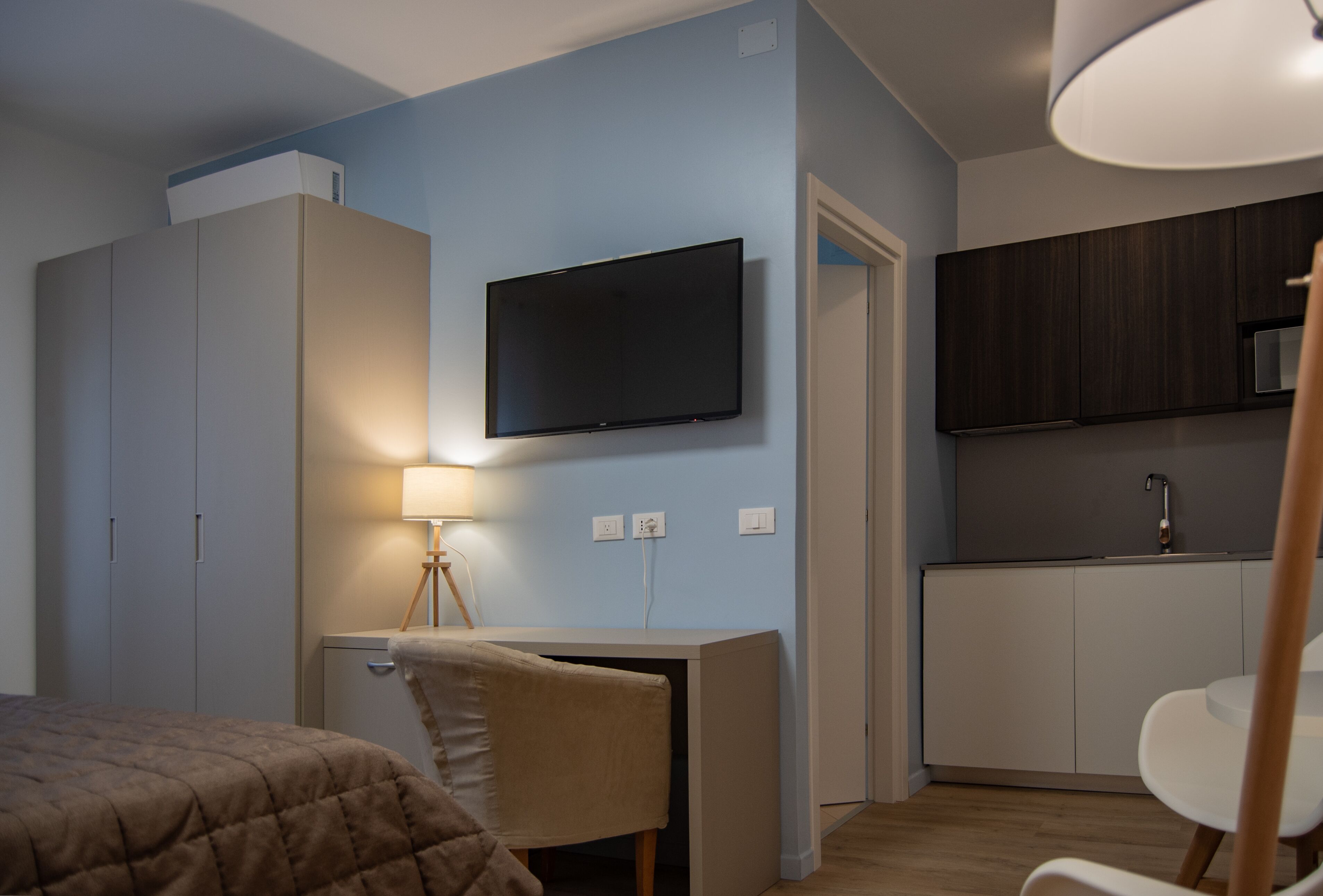 A compact room with a bed, a desk with a lamp, a TV, and a kitchenette.