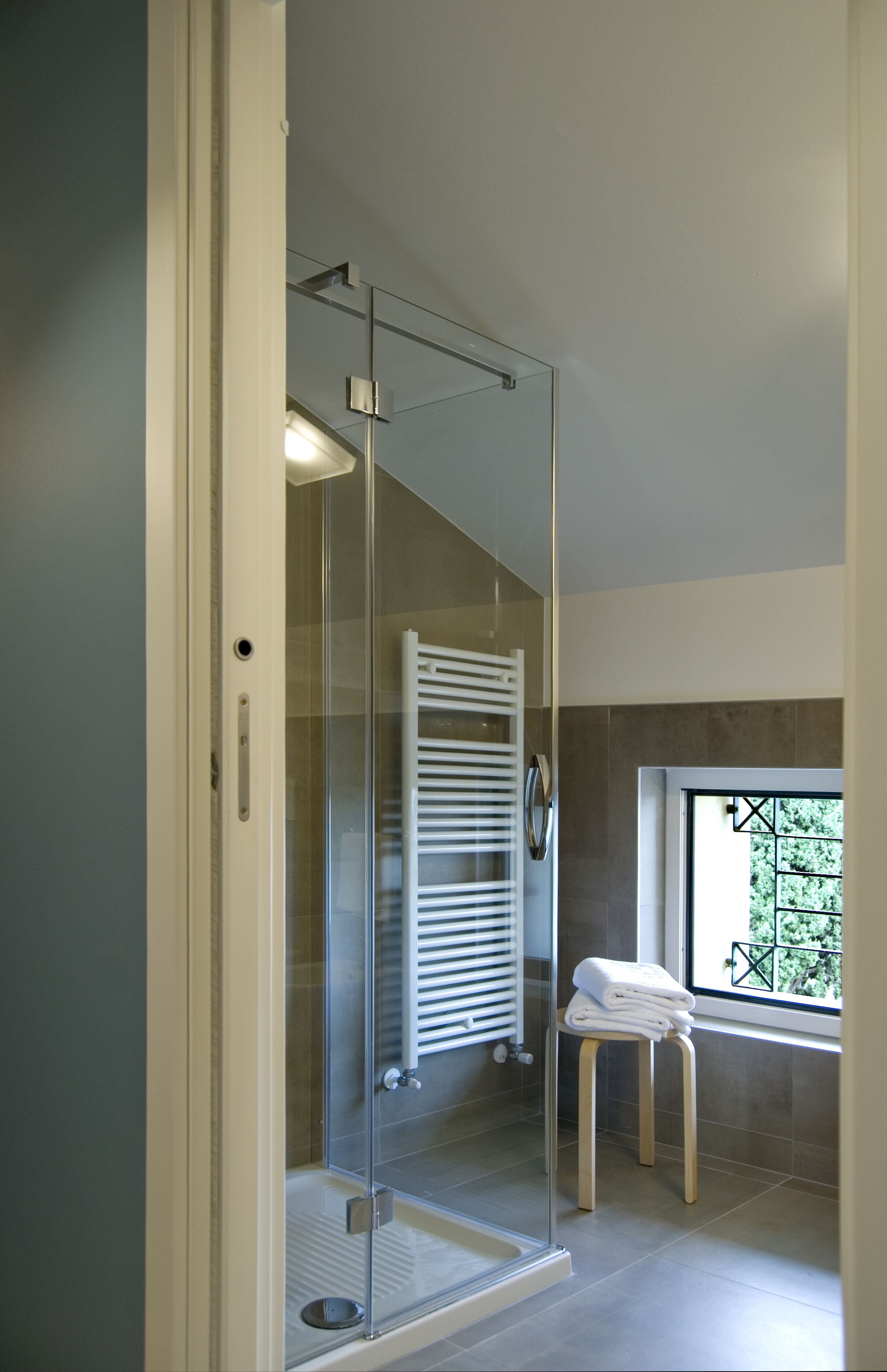 Un baño moderno con ducha de vidrio, una toalla blanca sobre un taburete y vistas al exterior con vegetación.