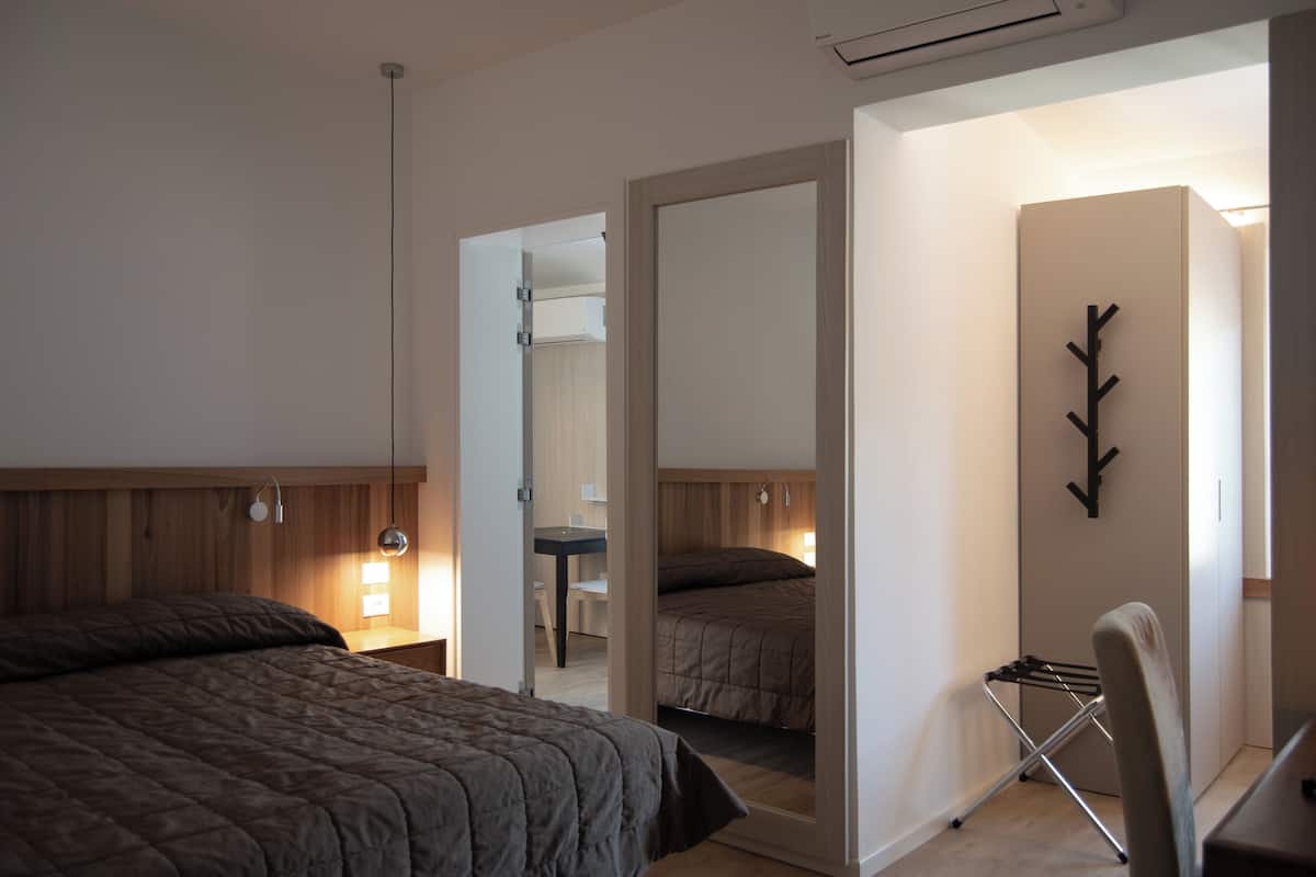 A bedroom with a large bed, a desk, and a chair. There is a mirror on the wall and a coat rack.