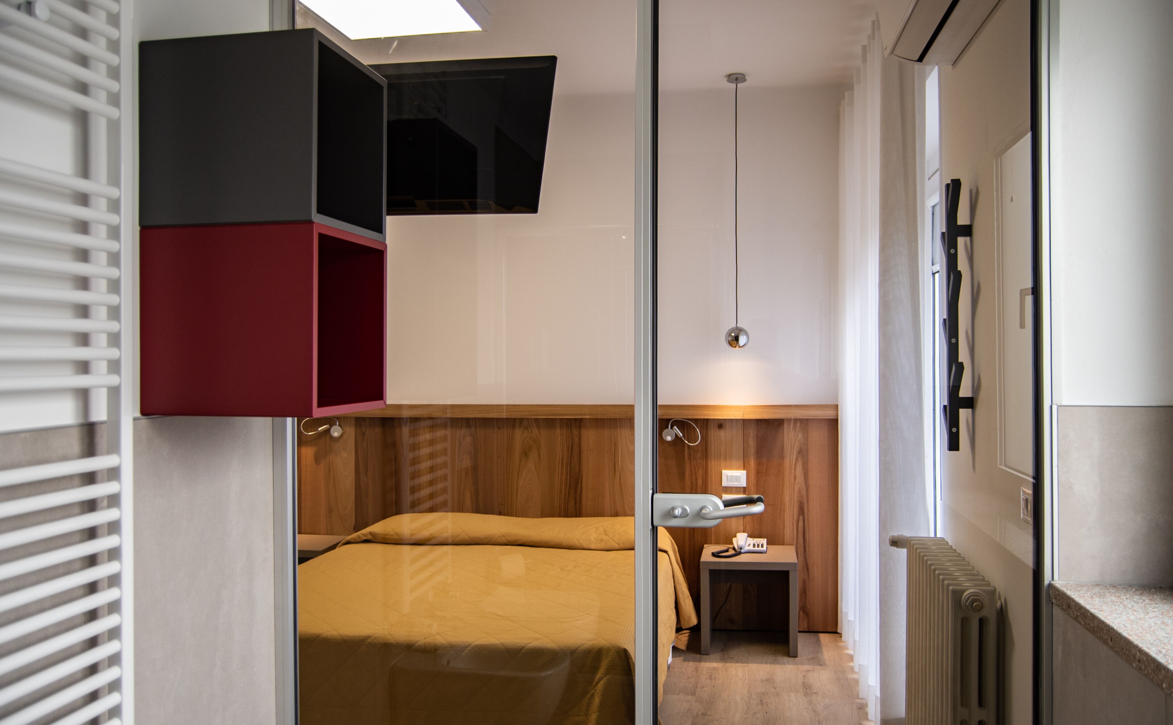 A bedroom with a bed, a small bedside table, a red and black cabinet, a wooden headboard, and a window with blinds.