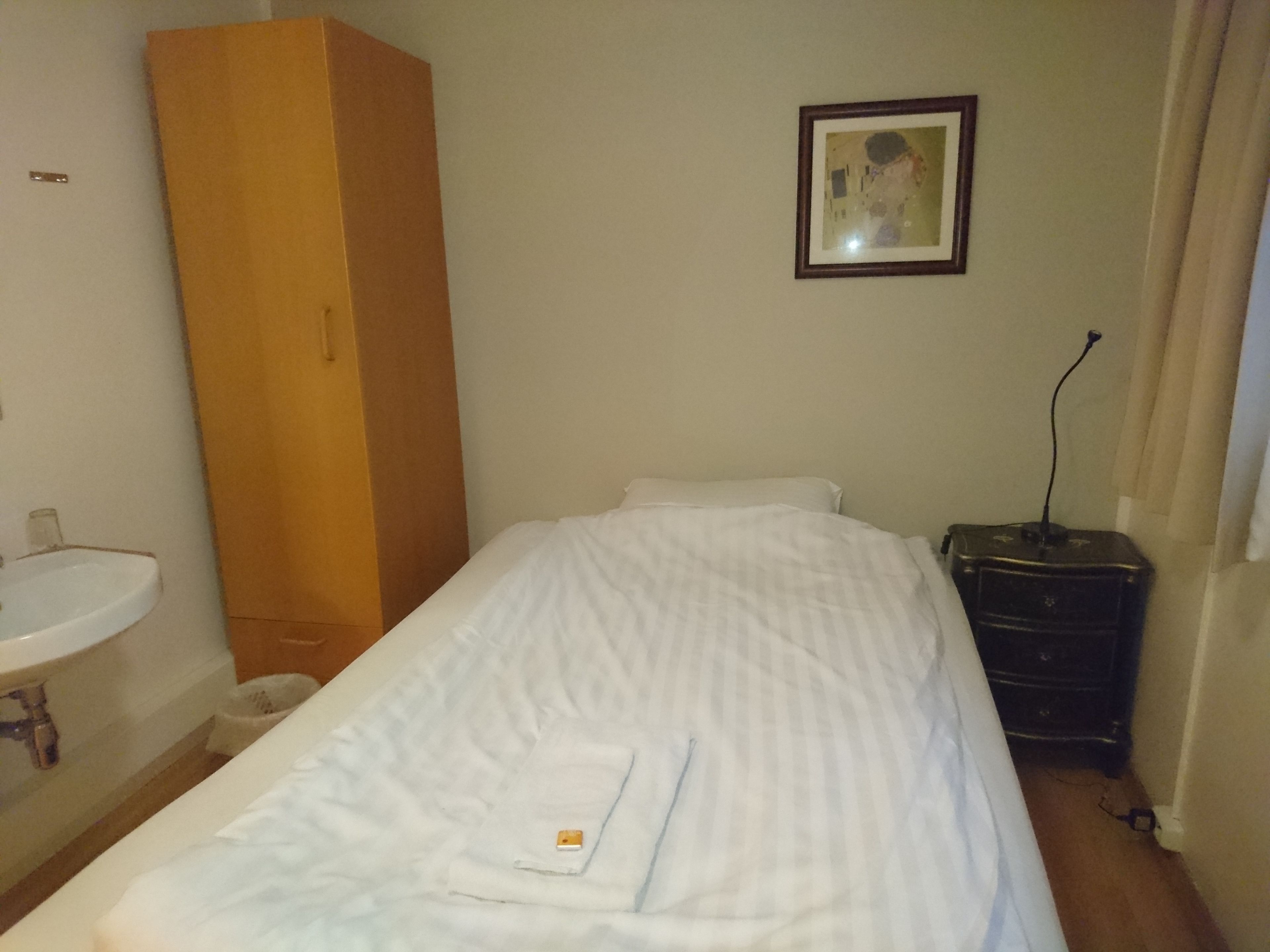 A small, single-bed room with a wooden wardrobe, a bedside table, a framed picture on the wall, and a bathroom with a sink and mirror.