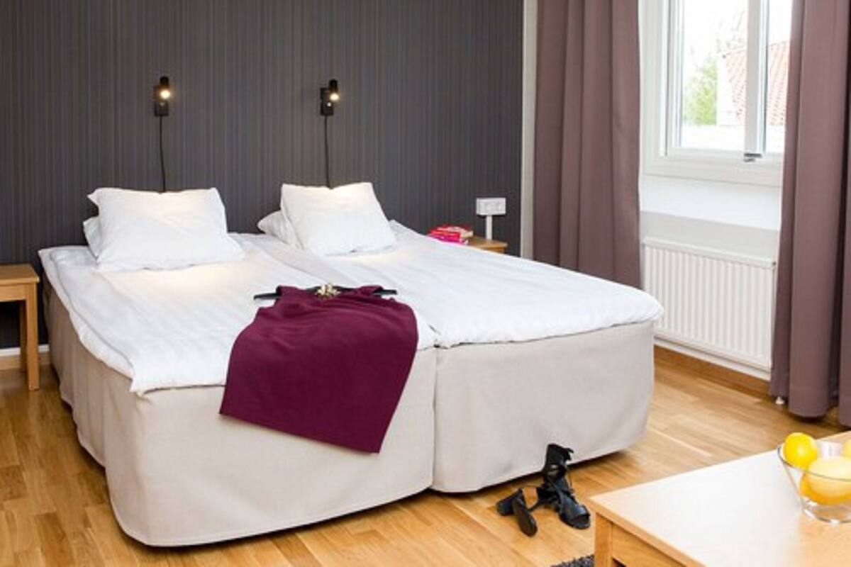 Una cama doble con ropa blanca de cama, un plaid morado y un par de zapatos negros en el suelo.