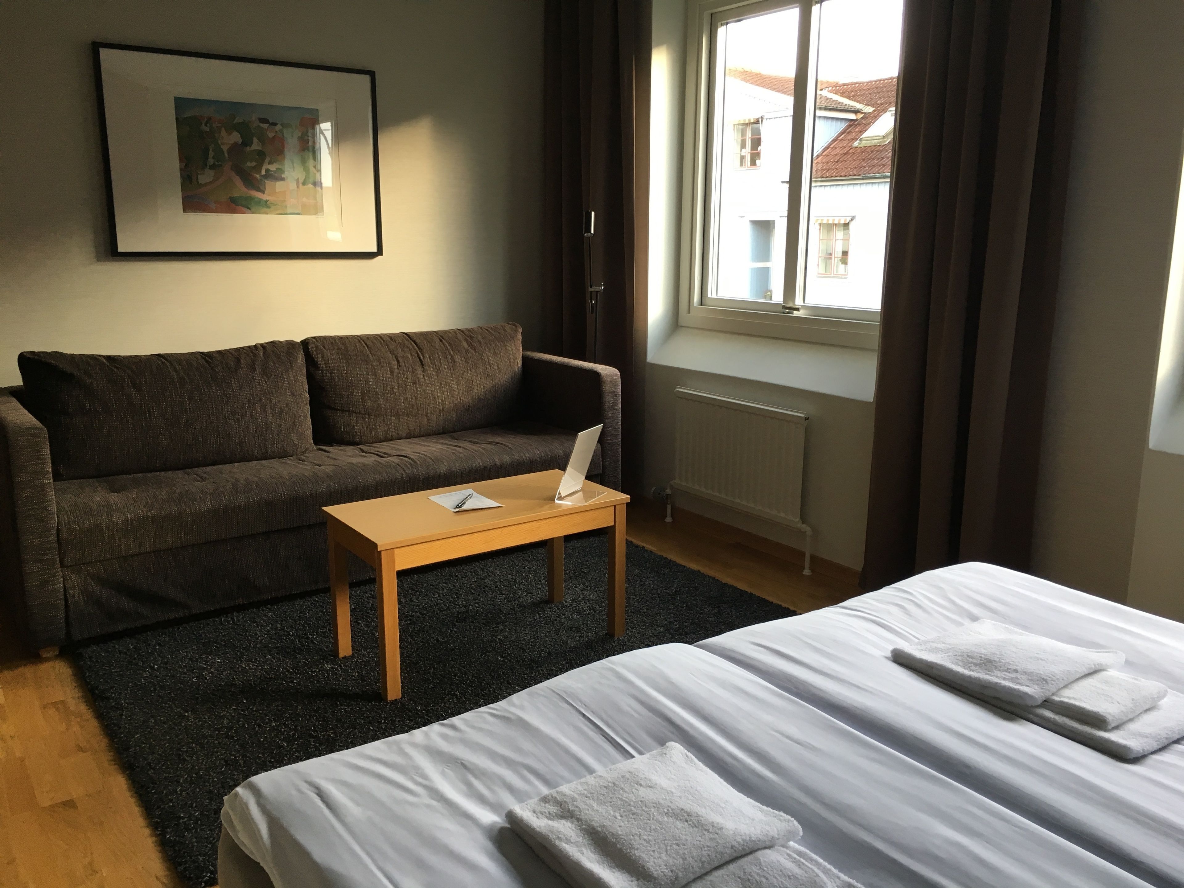 Une chambre d’hôtel avec un lit, un canapé, une petite table, un radiateur et une fenêtre avec des rideaux.