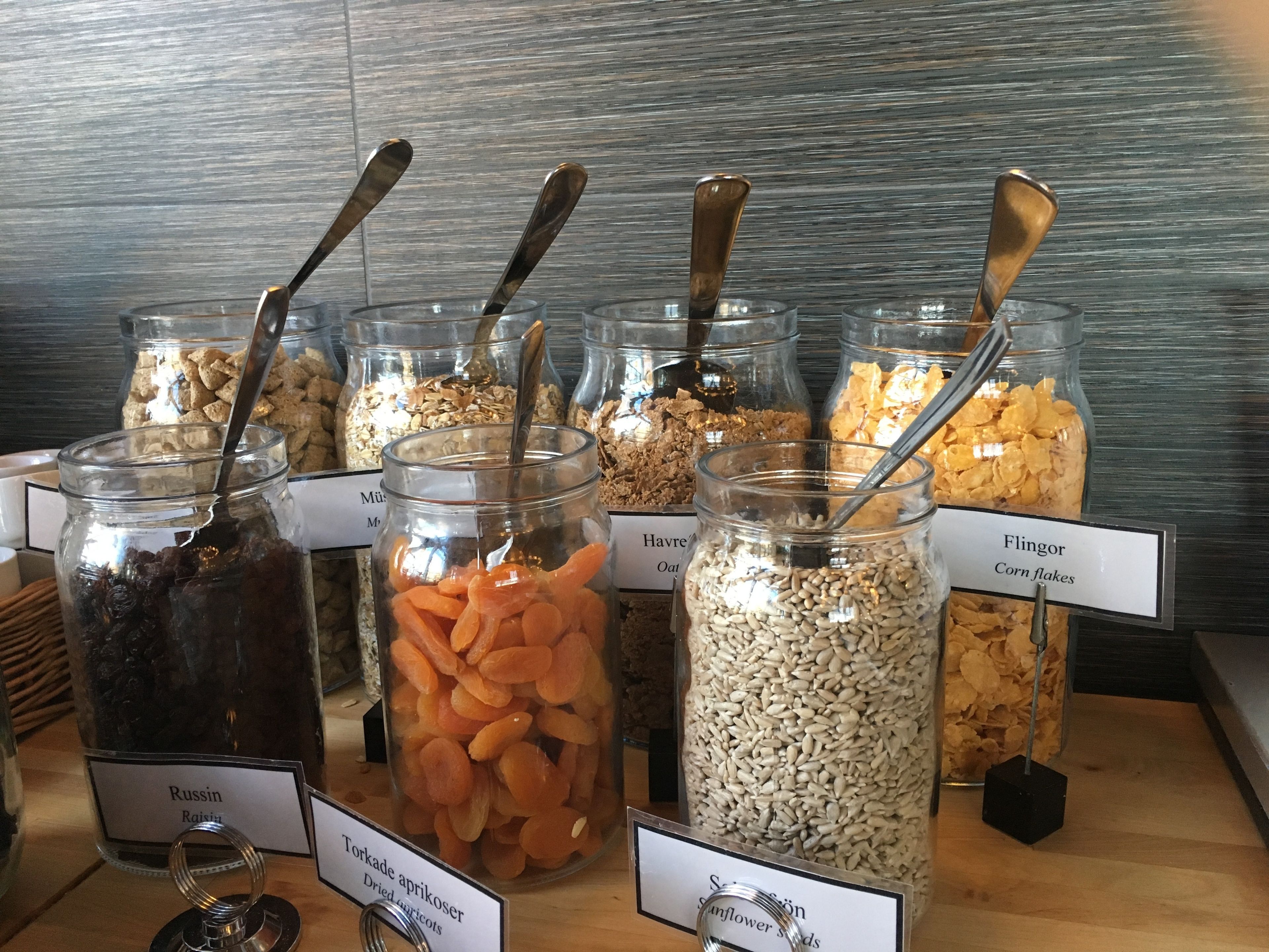 Verschiedene Lebensmittel in Gläsern, darunter Löffel, beschriftet mit Bezeichnungen wie Rosinen, getrocknete Aprikosen, Sonnenblumenkerne und Corn Flakes.