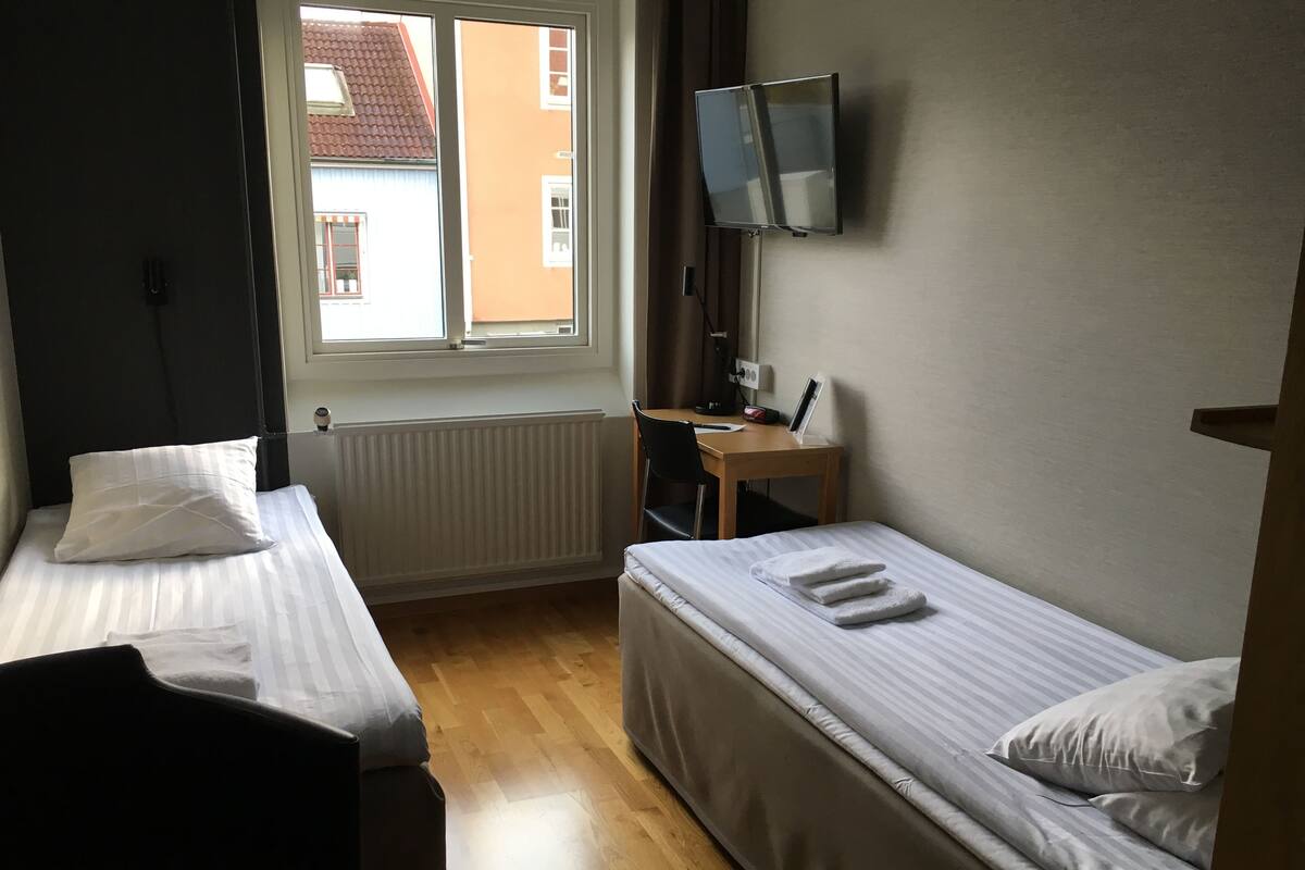 Una habitación pequeña y limpia con dos camas individuales, un escritorio con silla, un televisor montado en la pared y una ventana con vistas a edificios.