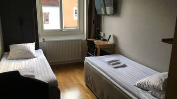 Una habitación pequeña y limpia con dos camas individuales, un escritorio con silla, un televisor montado en la pared y una ventana con vistas a edificios.