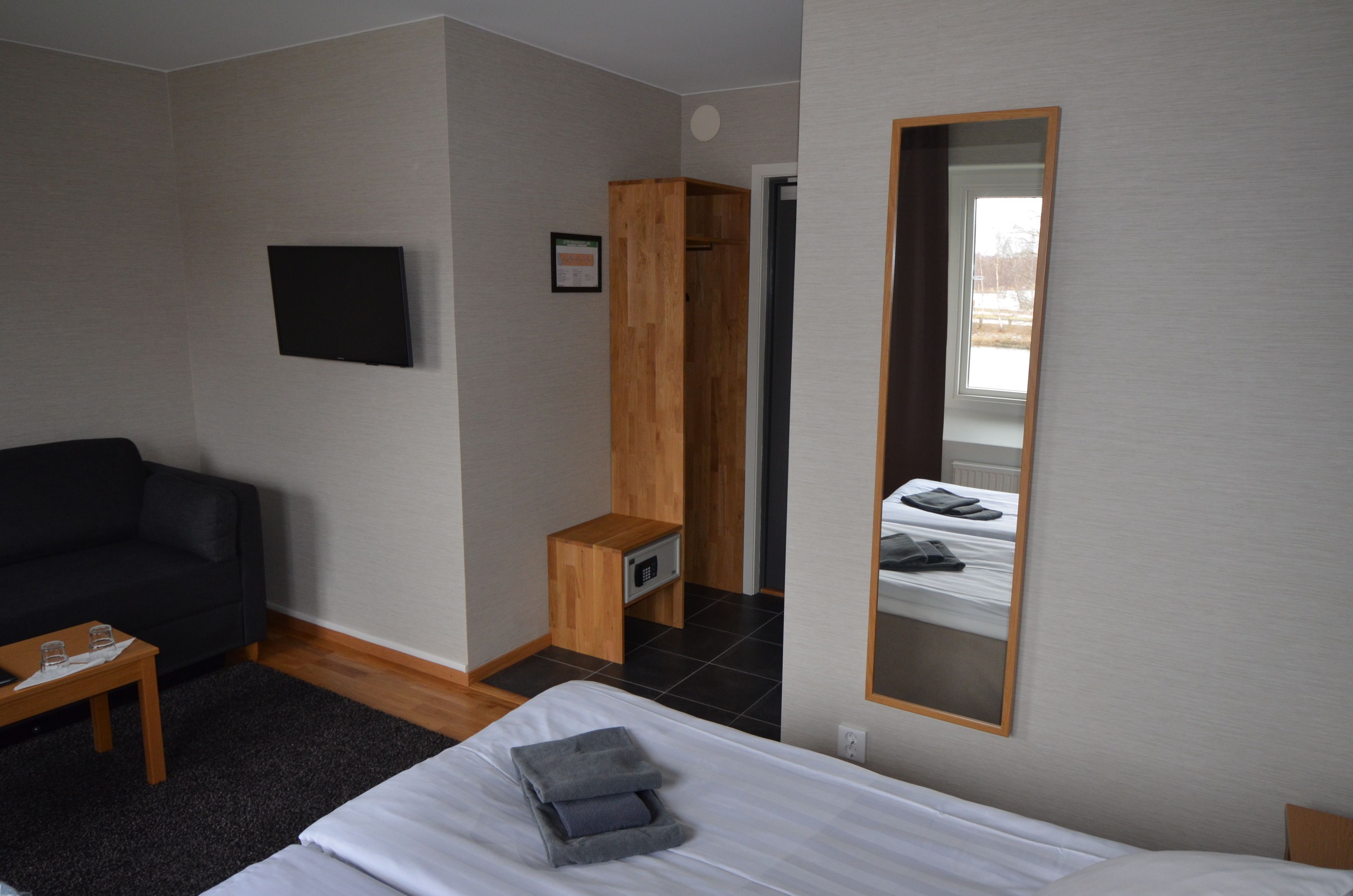A bedroom with a bed, a wooden wardrobe, a small table, a black sofa, and a TV mounted on the wall.