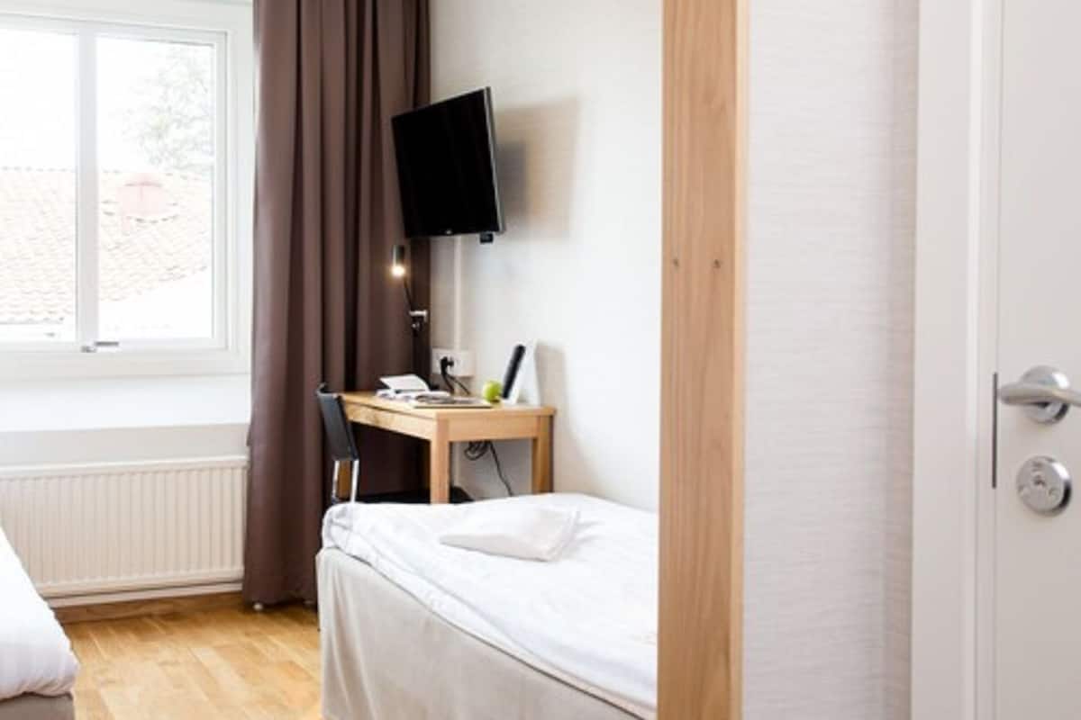 Un dormitorio con cama, escritorio con computadora, silla, televisor en la pared y ventana con cortinas.