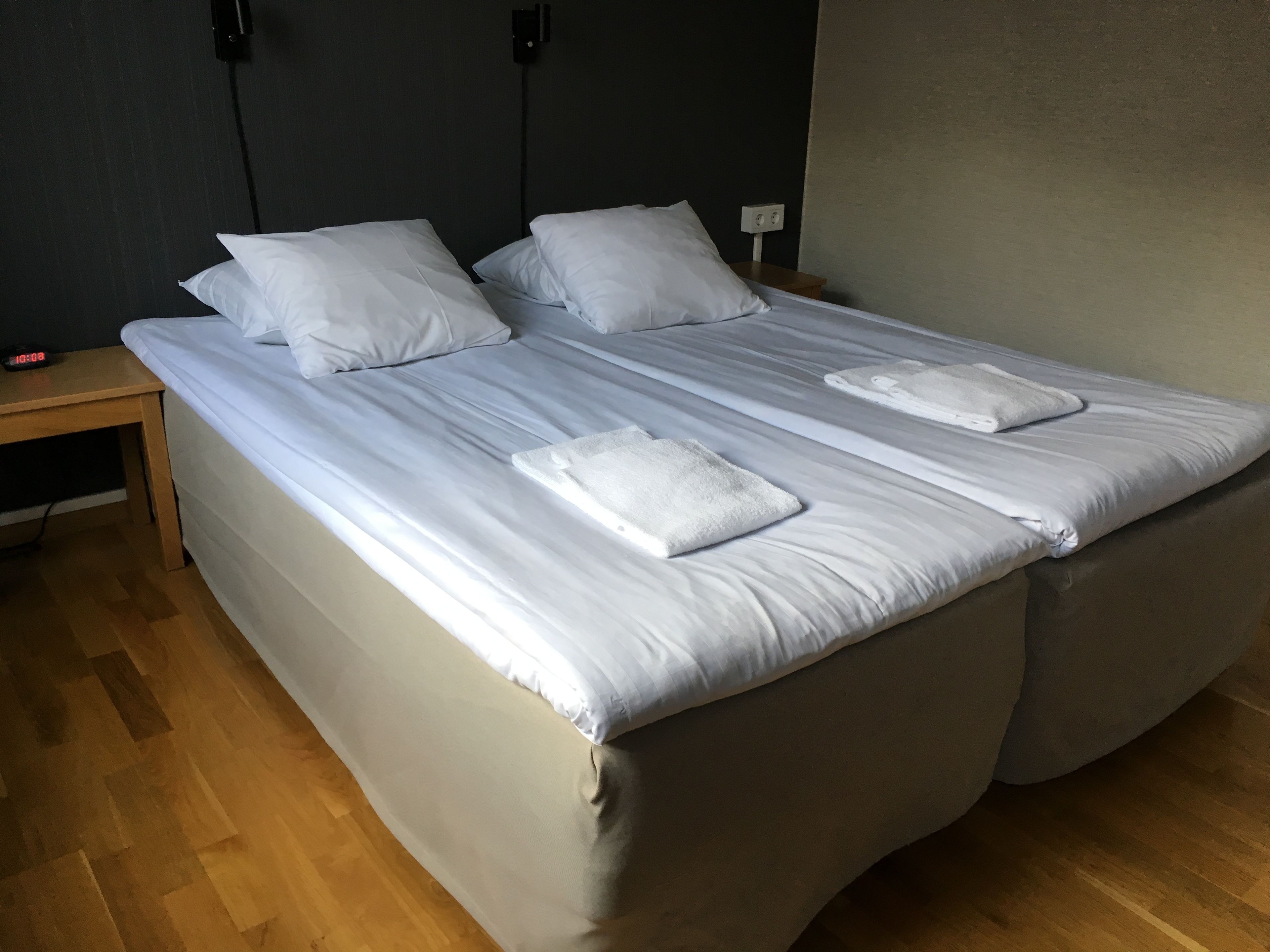 A neatly made bed with white linens and two towels, a wooden nightstand with a clock, and a plain wall.