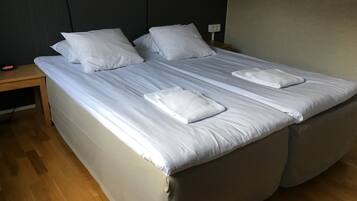 A neatly made bed with white linens and two towels, a wooden nightstand with a clock, and a plain wall.