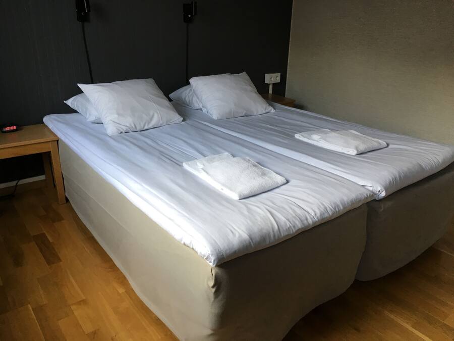 A neatly made bed with white linens and two towels, a wooden nightstand with a clock, and a plain wall.