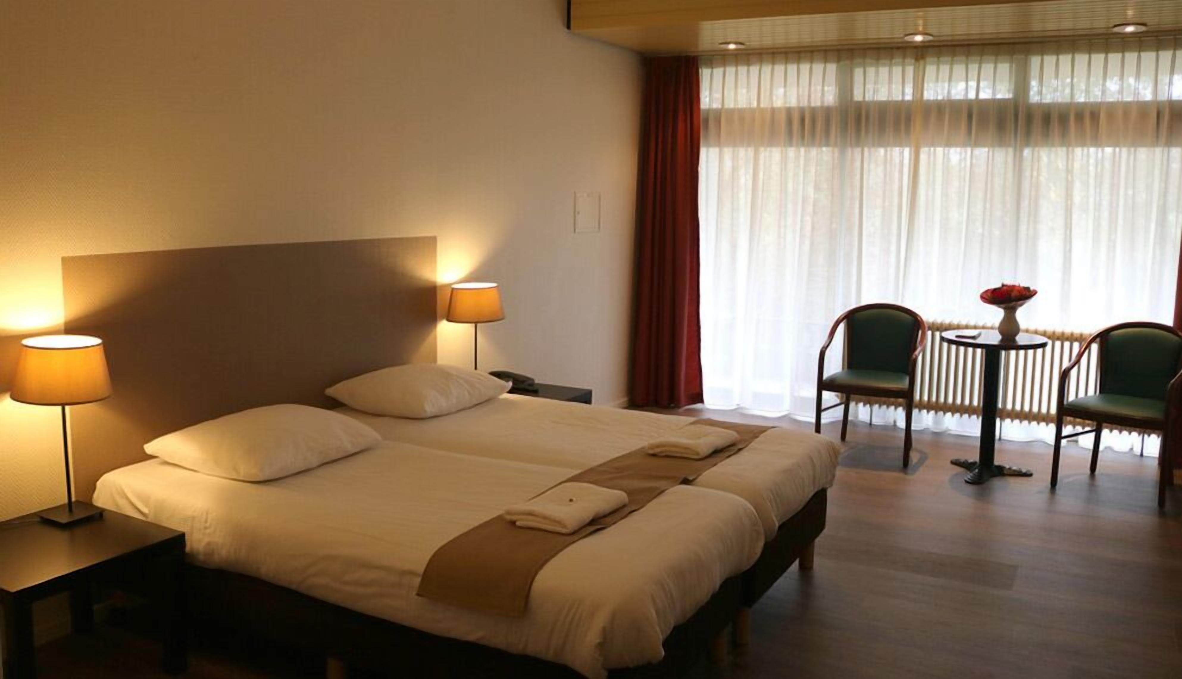 Une chambre d’hôtel avec un grand lit, deux tables de chevet avec des lampes, une petite table ronde avec deux chaises, et une vue à travers des rideaux légers.