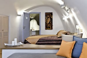 A bedroom with a bed, bedside table, lamp, and a painting on the wall. - Anemomilos Hotel & Apartments (Santorini)