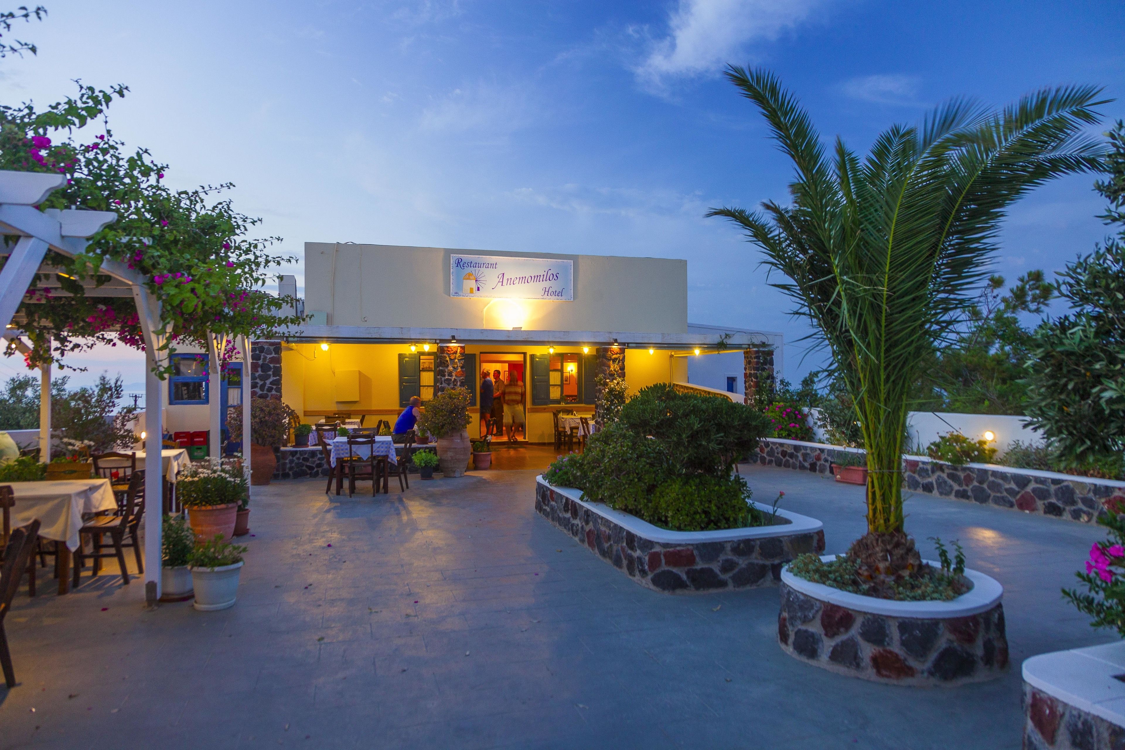 A restaurant with outdoor seating, a stone planter with a palm tree, and a sign for the Anemomilos Hotel.