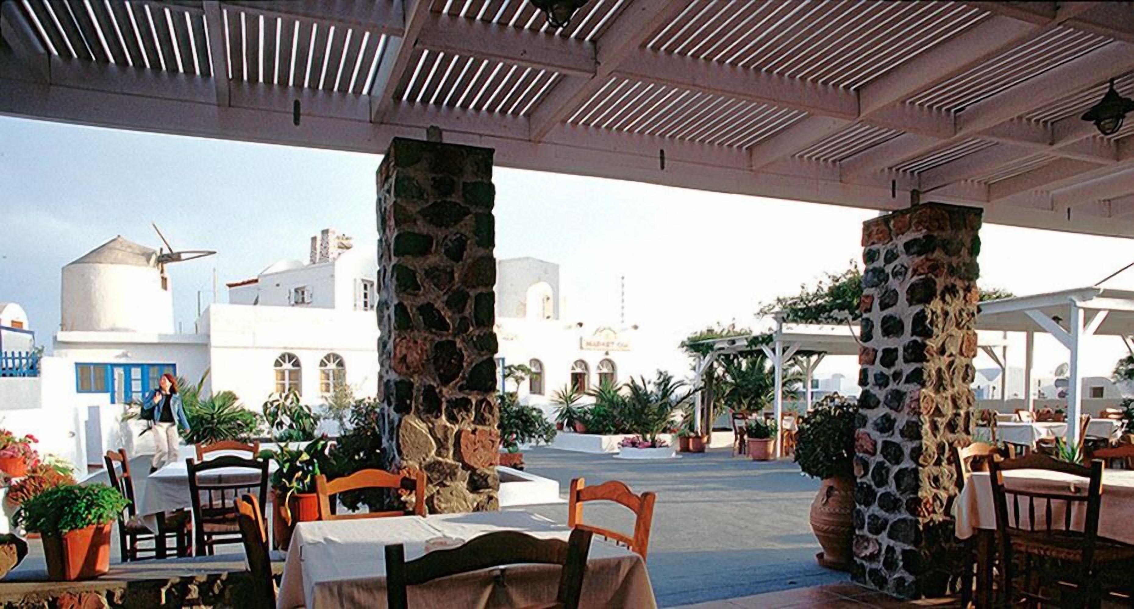 An outdoor seating area with stone pillars, tables, and chairs, overlooking a white building with blue accents and a windmill.