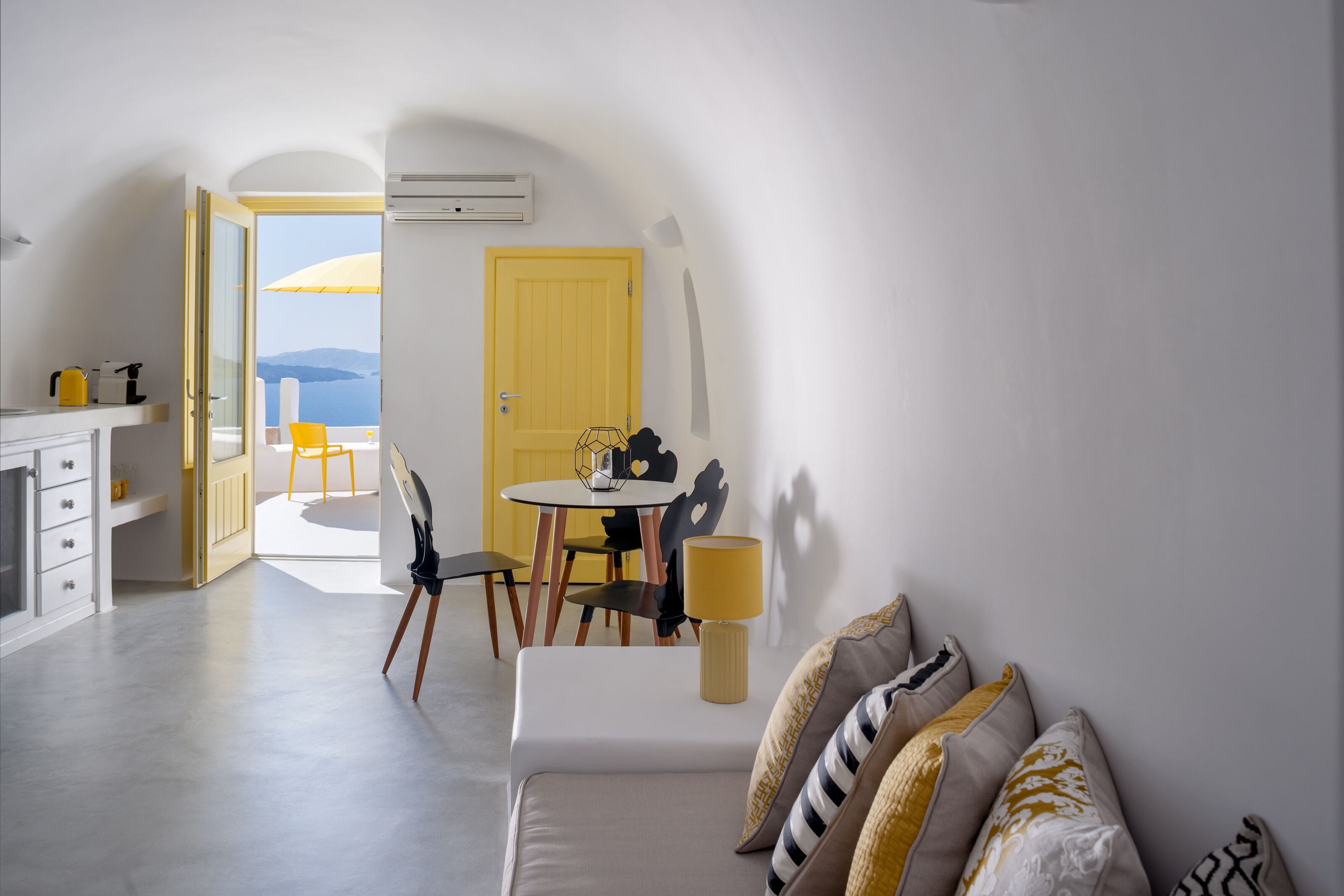 A modern, minimalist living room with a sofa, a dining table with chairs, and a view of the sea through an open door.