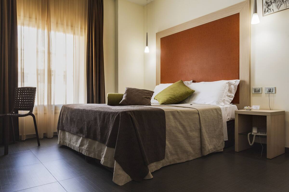 A hotel room with a bed, a chair, a nightstand, and a window with curtains.
