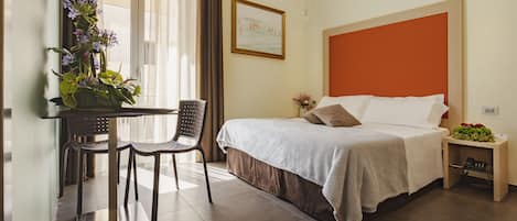 A hotel room with a bed, a table with chairs, a small bedside table with a vase of flowers, and a painting on the wall.