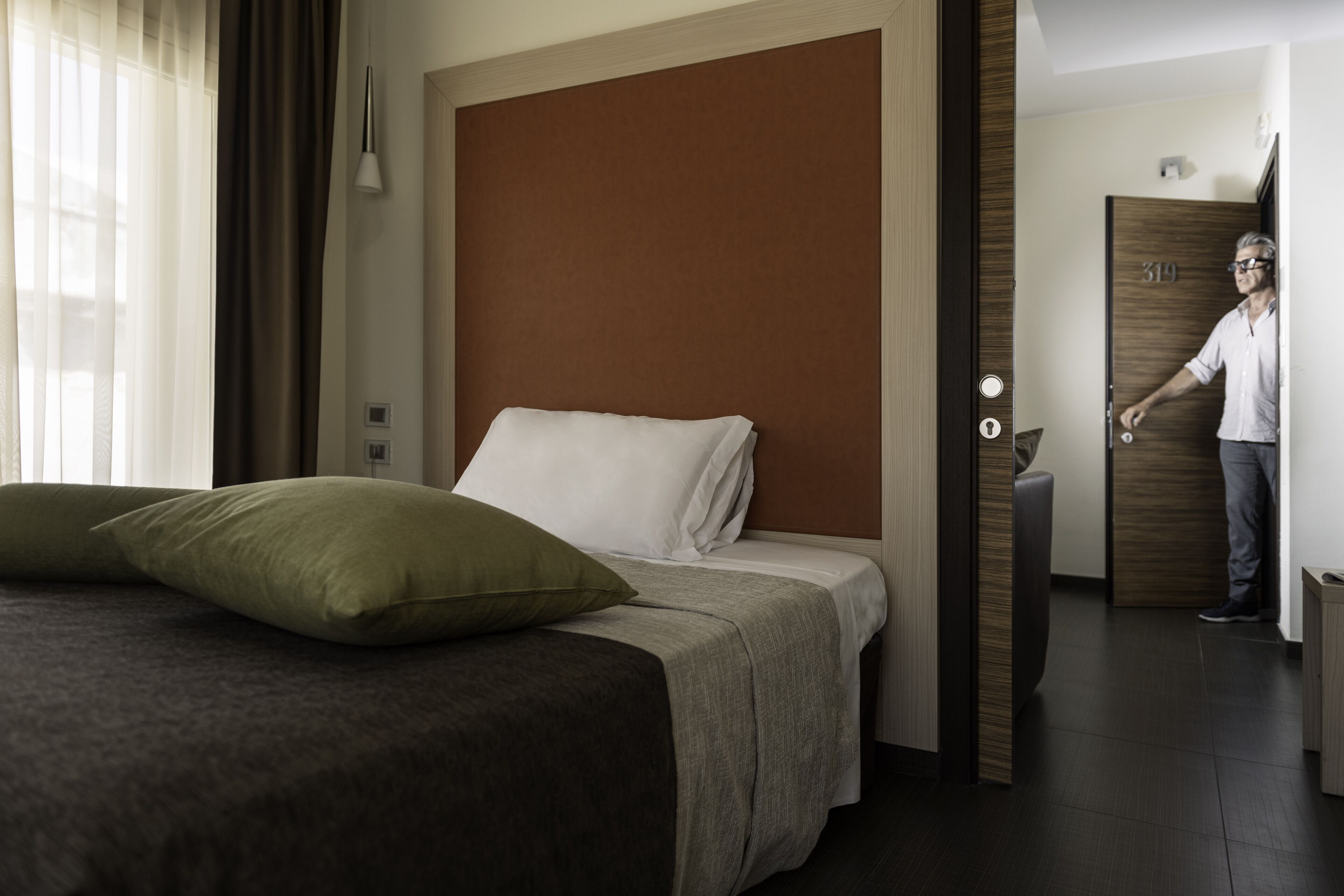 Una camera d'albergo moderna con un letto grande, una testiera in legno e una porta che dà su un corridoio.
