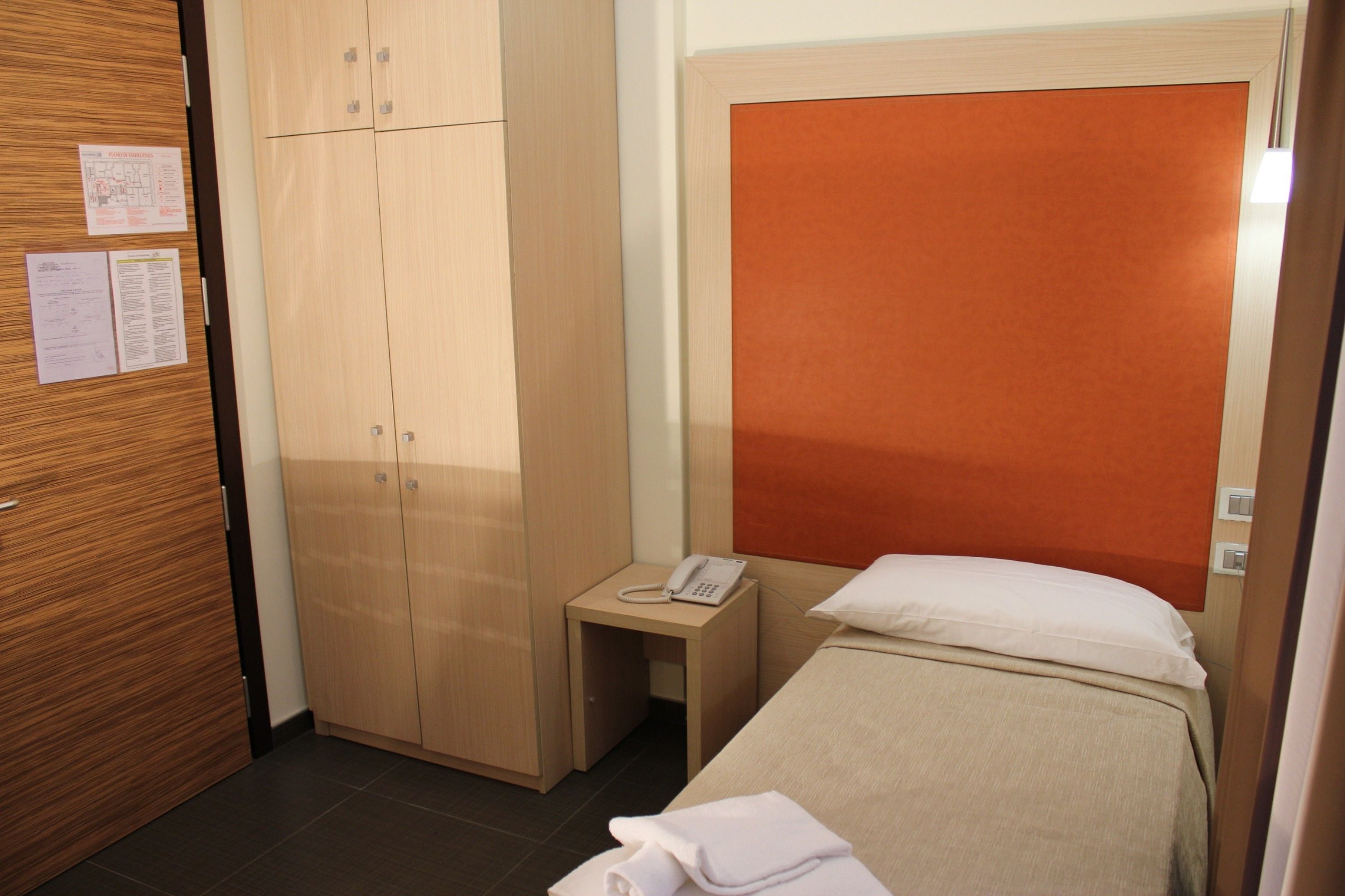 A hotel room with a bed, a wooden wardrobe, a small bedside table with a phone, and a wall-mounted bulletin board.